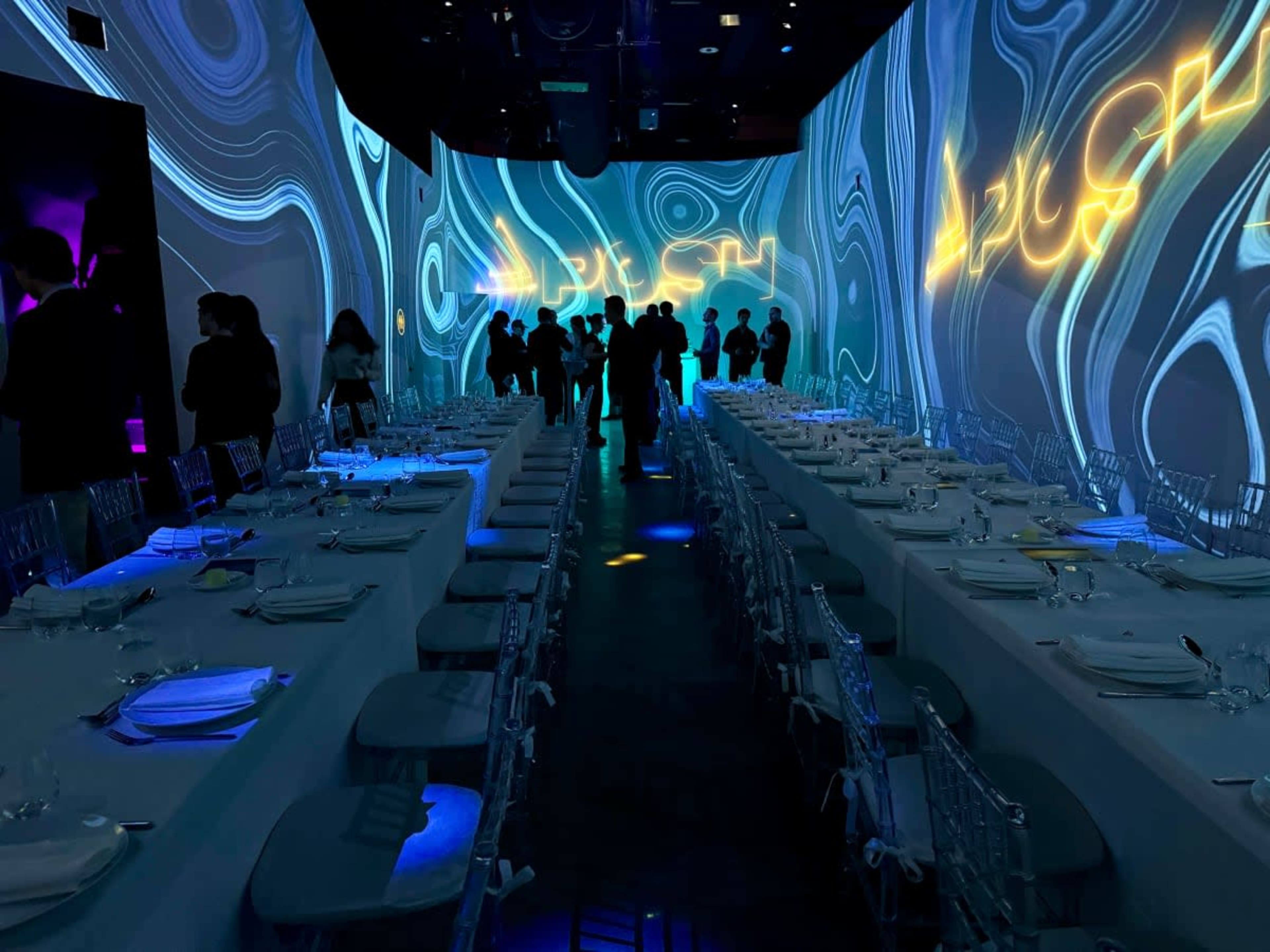A dimly lit dining hall features long tables set with elegant tableware, surrounded by guests silhouetted against colorful projected designs on the walls.