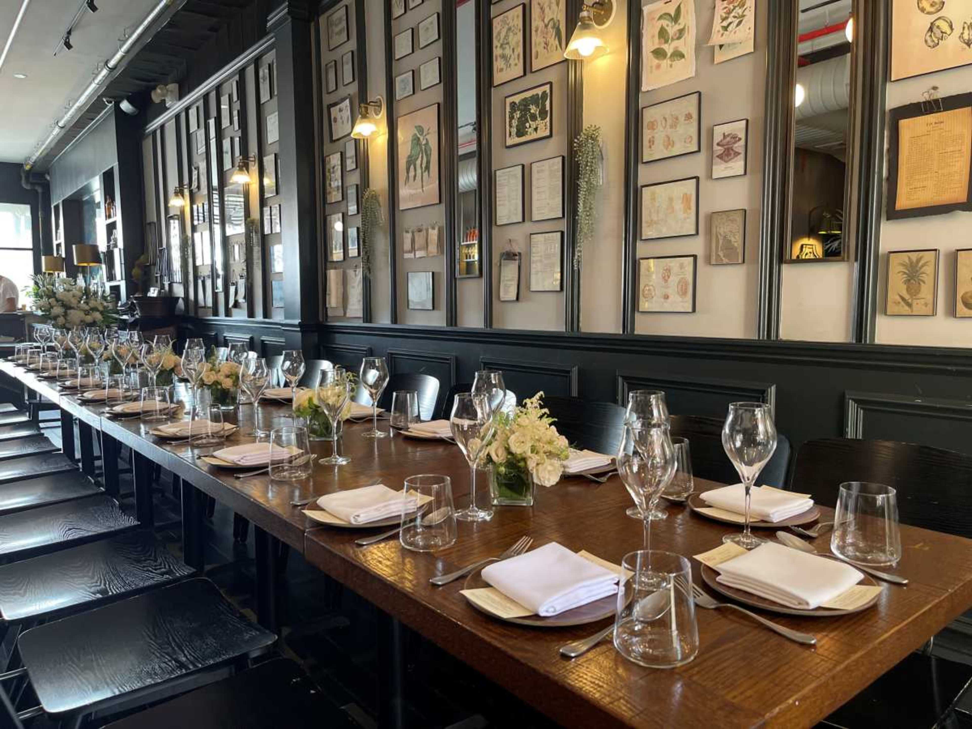 A long wooden table is set with glassware and white plates, surrounded by black chairs in a restaurant adorned with framed botanical illustrations on the walls.