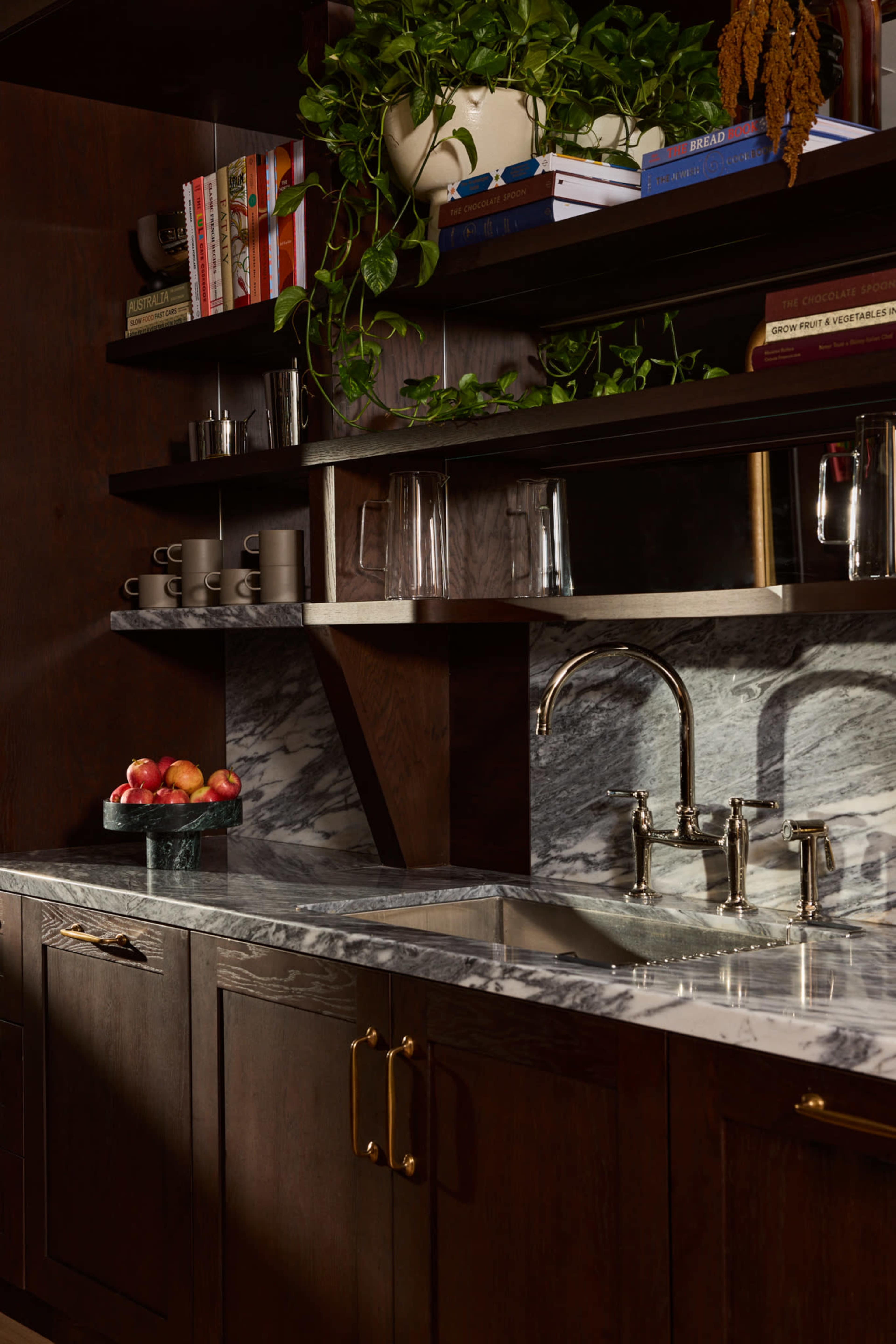 A modern kitchen features dark wooden cabinetry, a marble countertop with a sink, and shelves adorned with plants, books, and coffee mugs.