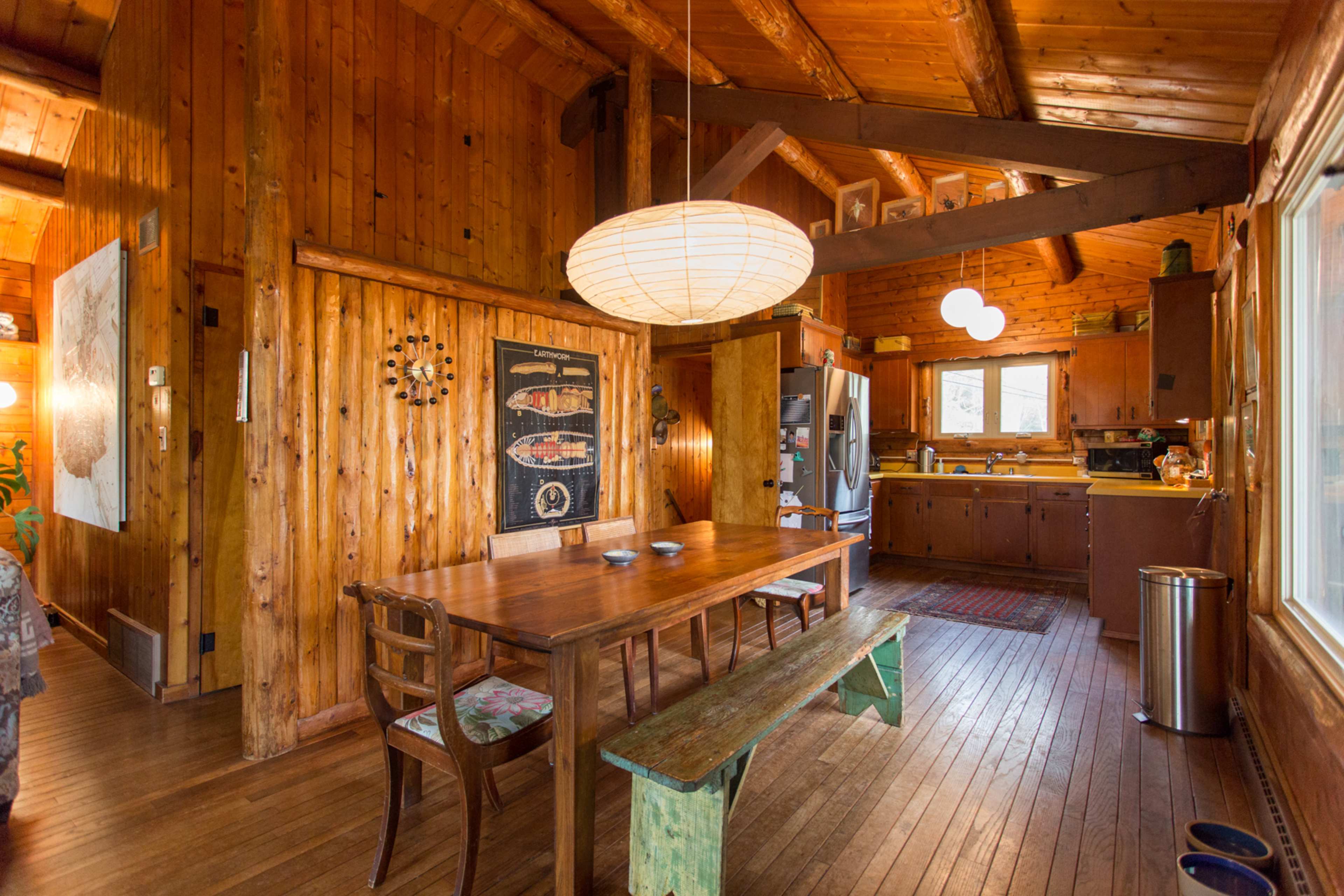 The scene features a wooden dining area with a large table, surrounded by chairs, next to a kitchen with a window and hanging light fixtures.