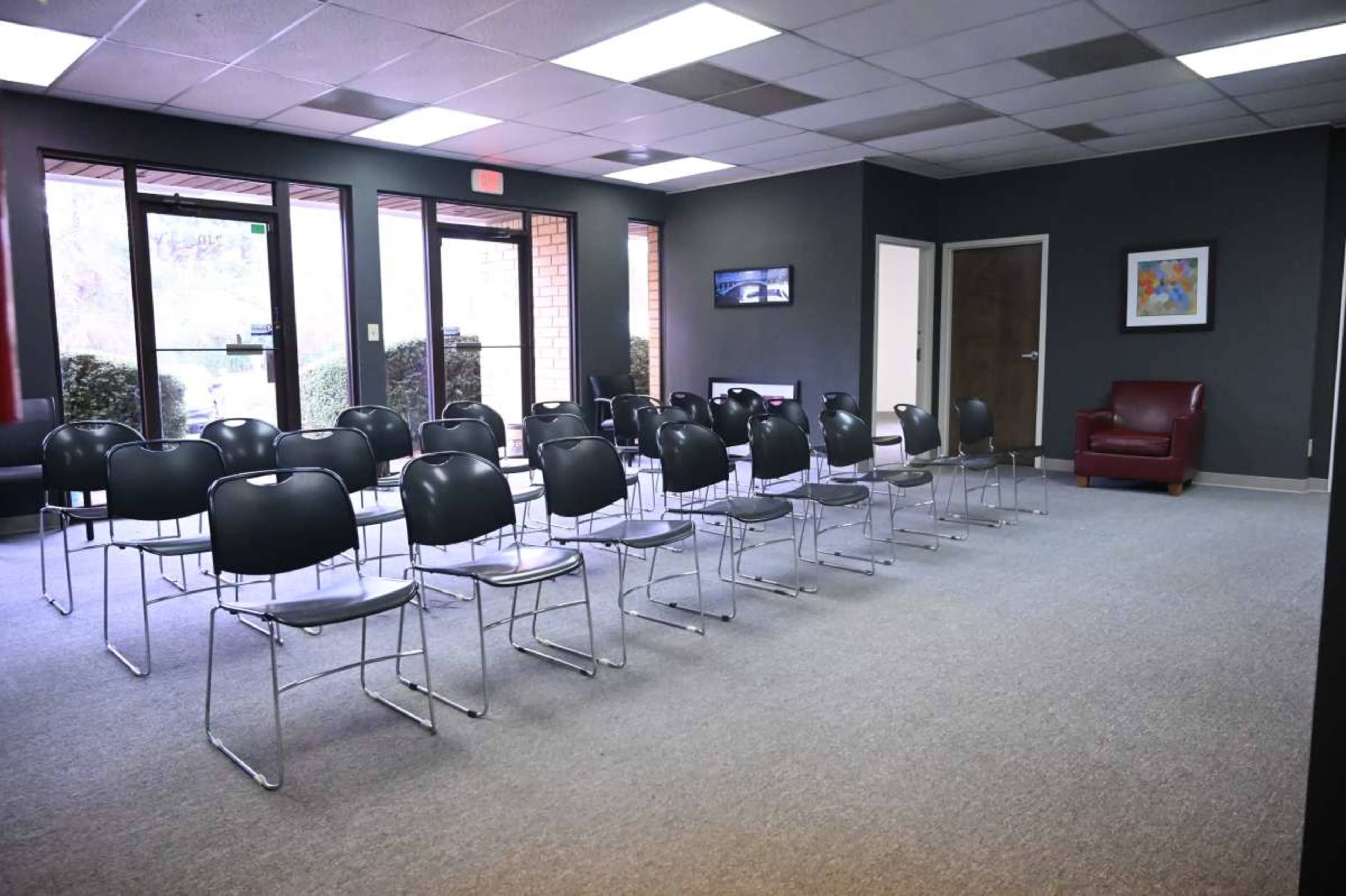 The image shows a waiting room with several empty black chairs arranged in rows and a red armchair in the corner.