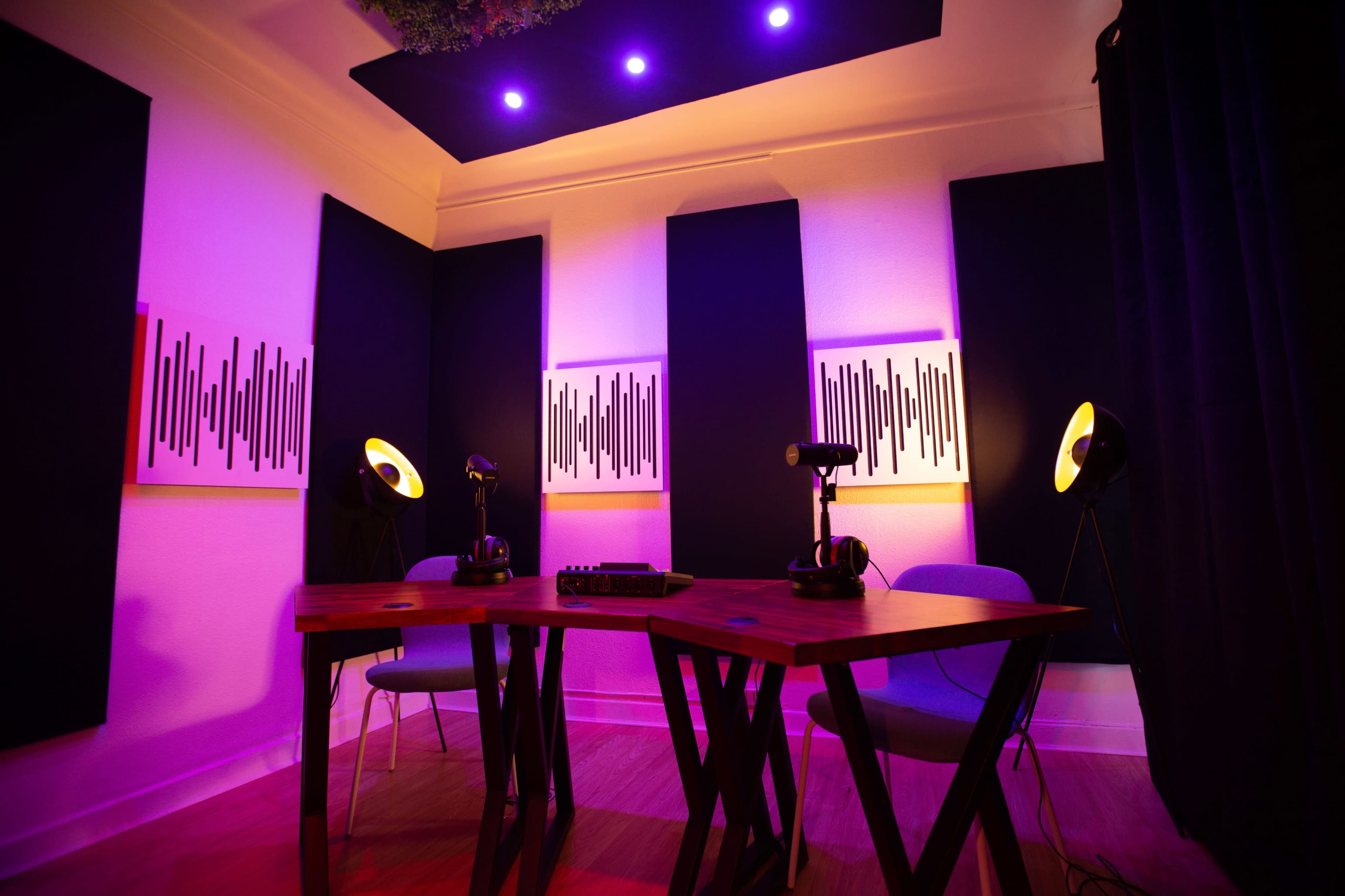 A small recording studio with a wooden table, two microphone setups, and wall art featuring sound wave patterns, illuminated by purple lights.