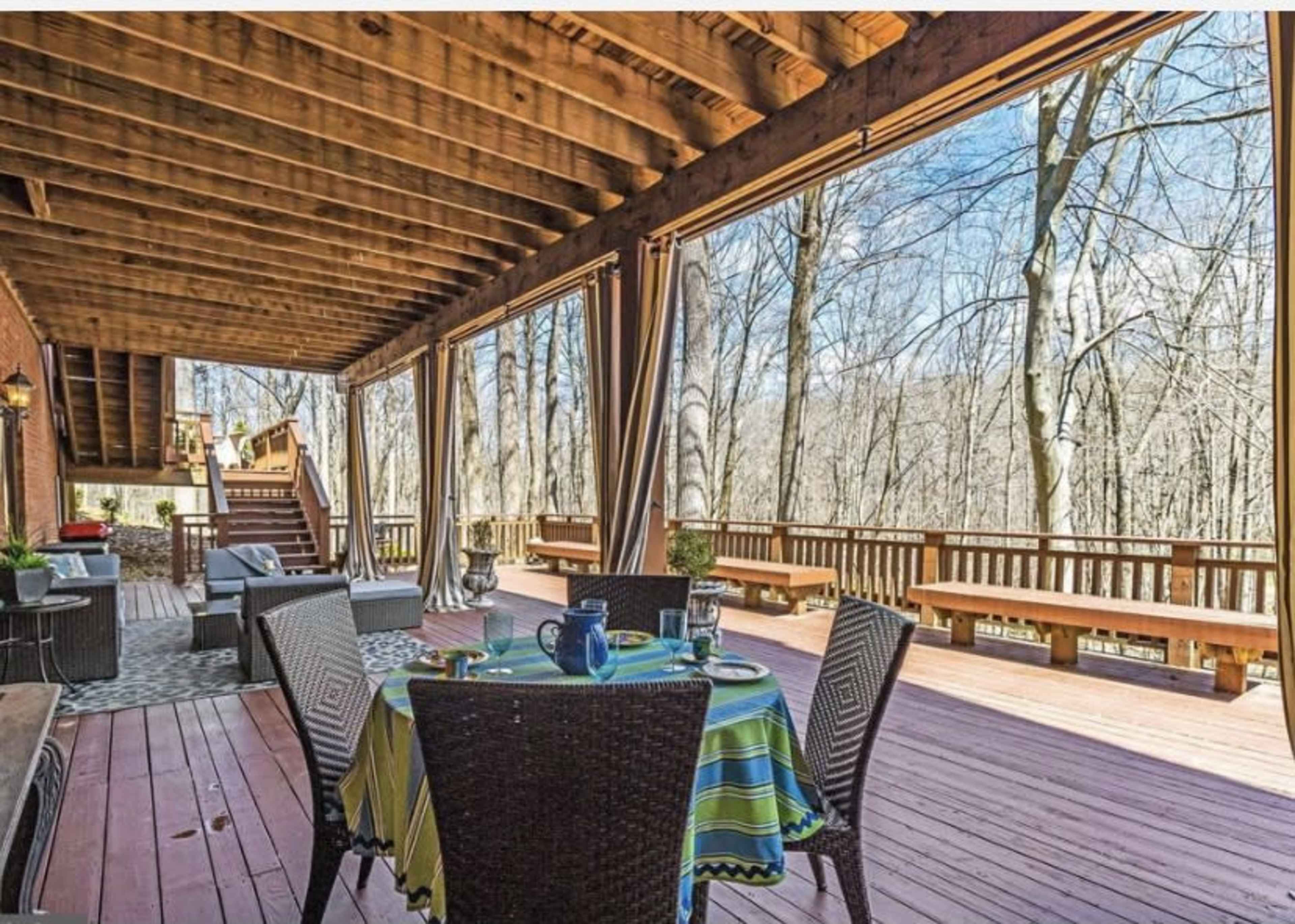 The image shows a spacious wooden deck with outdoor seating, a dining table set for breakfast, and large windows overlooking a wooded area.