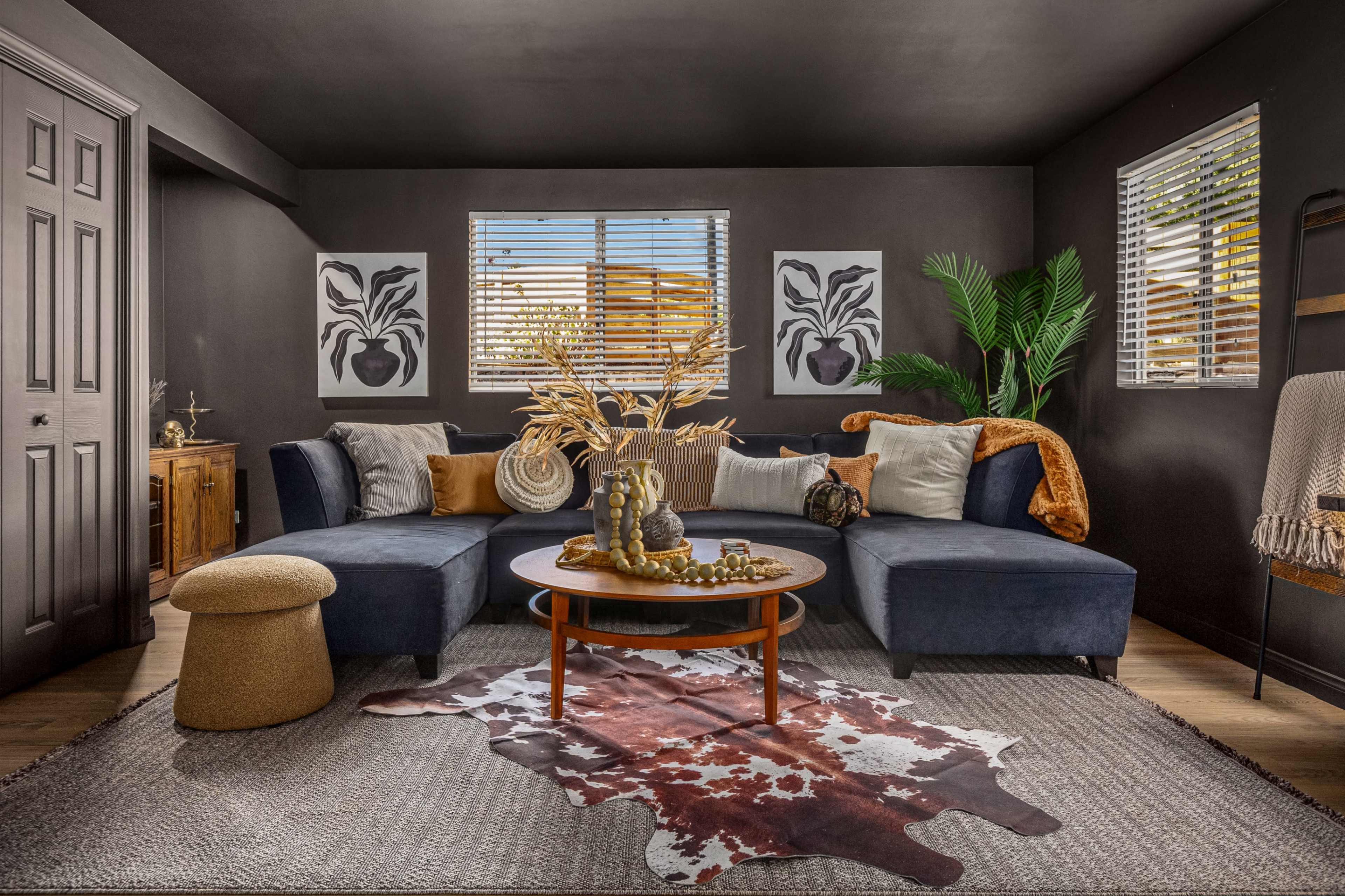 The image depicts a modern living room featuring a large sectional sofa, a round coffee table, and decorative elements including plants and artwork on the walls.