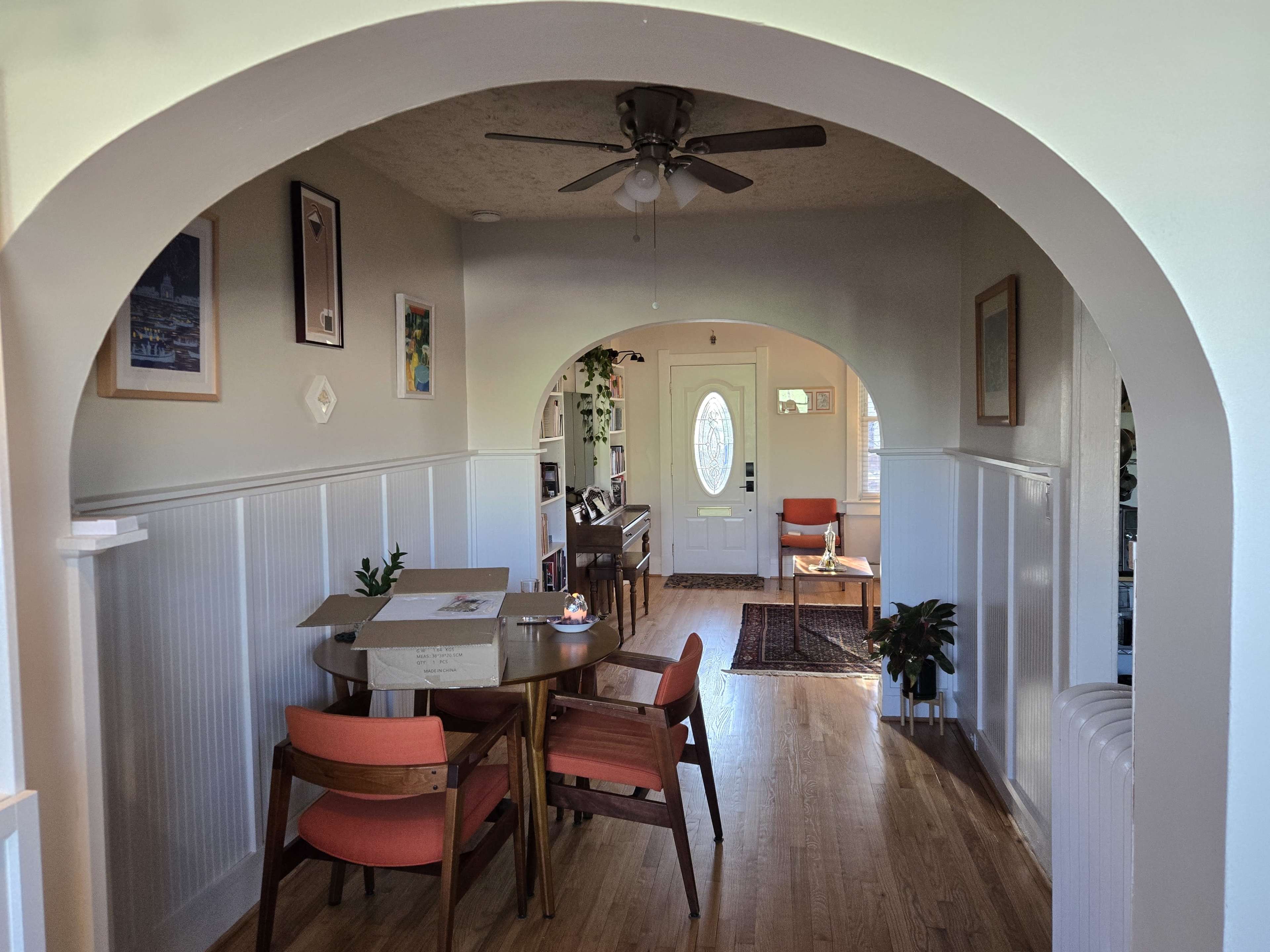 The image shows a well-lit hallway leading from a dining area with a table and chairs to a living room with a visible front door and various pieces of furniture.