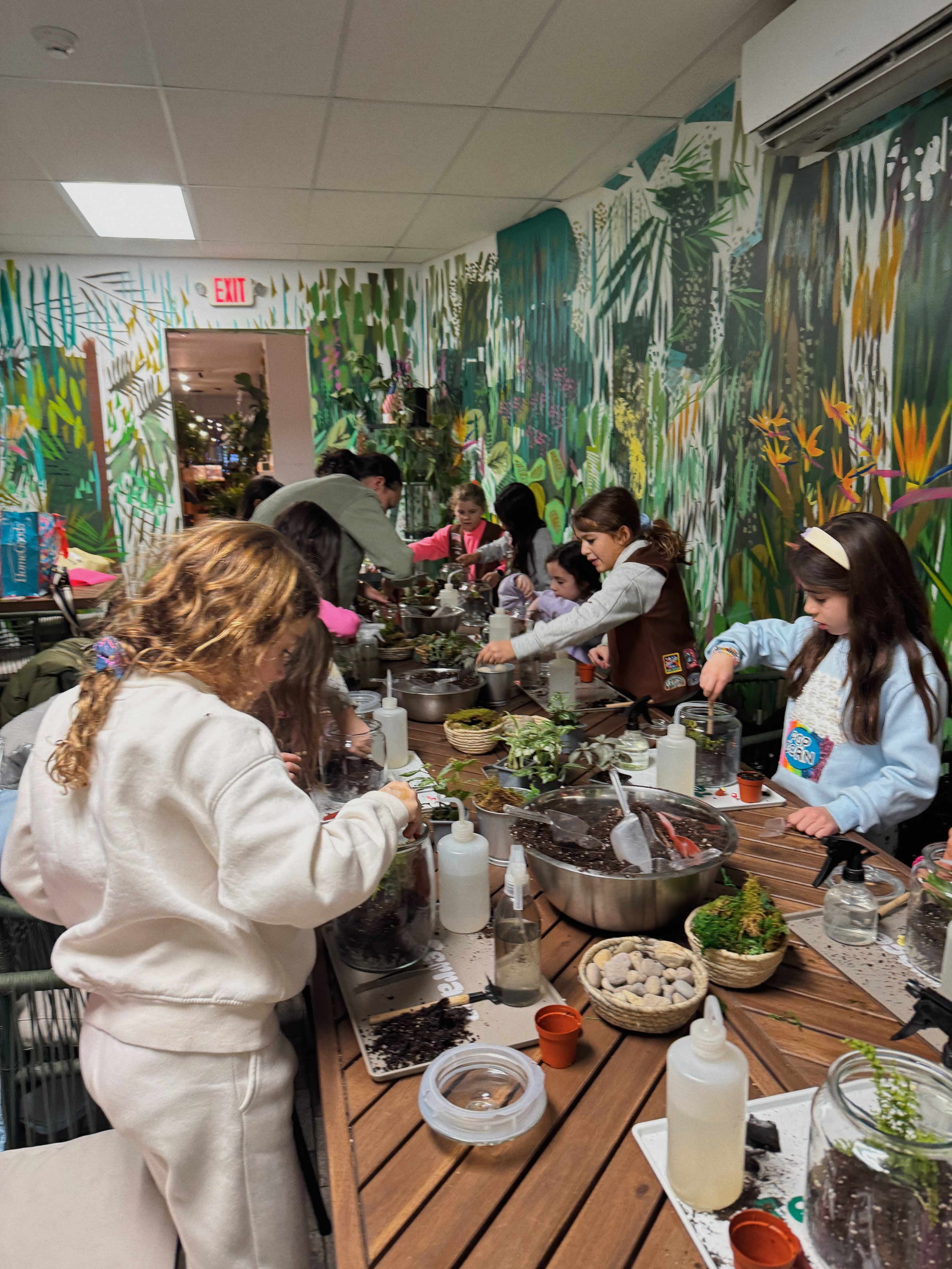 Children are engaged in a hands-on activity creating plant arrangements at a long wooden table surrounded by tropical-themed murals.