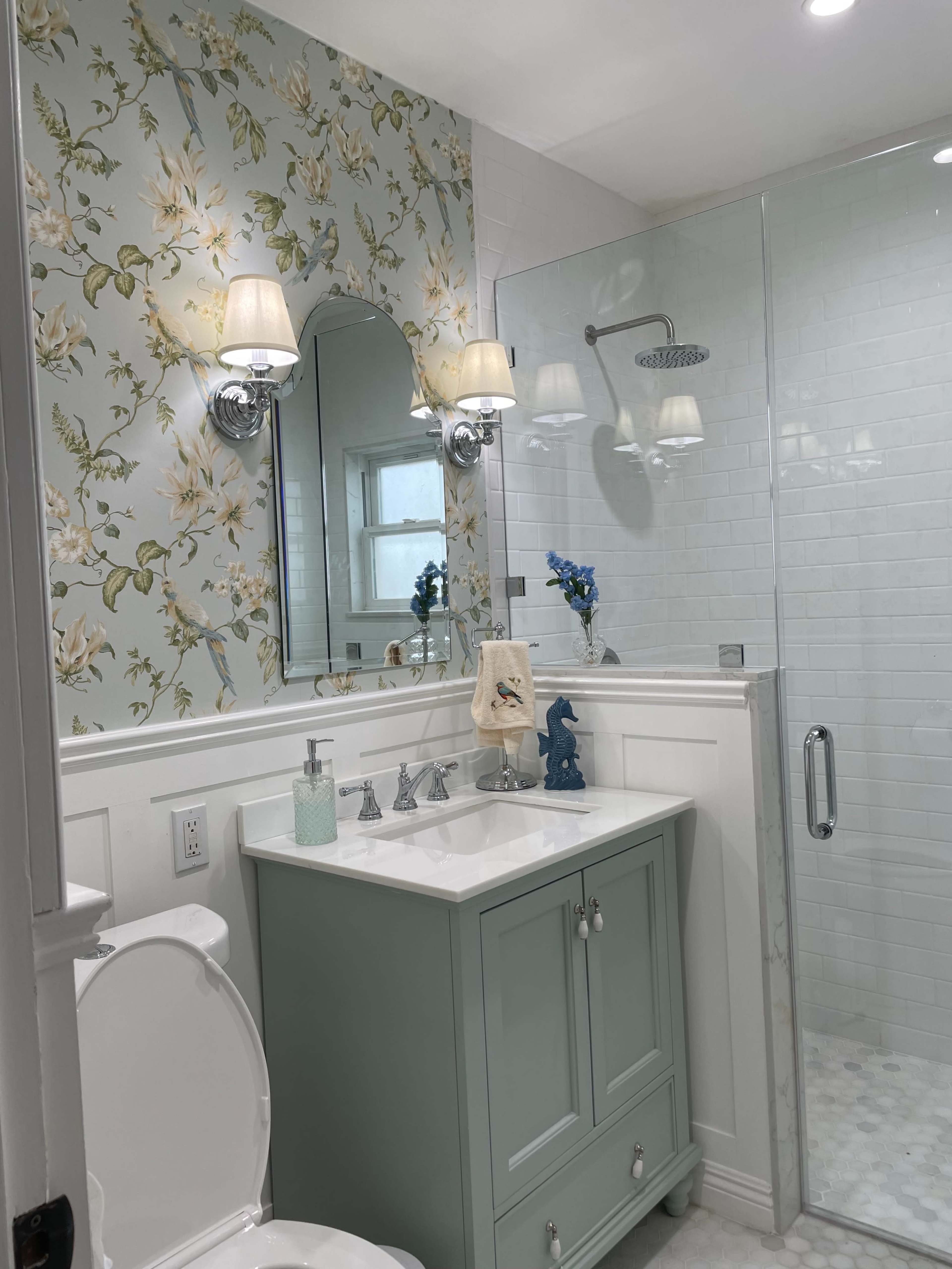 A bathroom featuring a light blue vanity, floral wallpaper, a glass shower enclosure, and a white toilet.