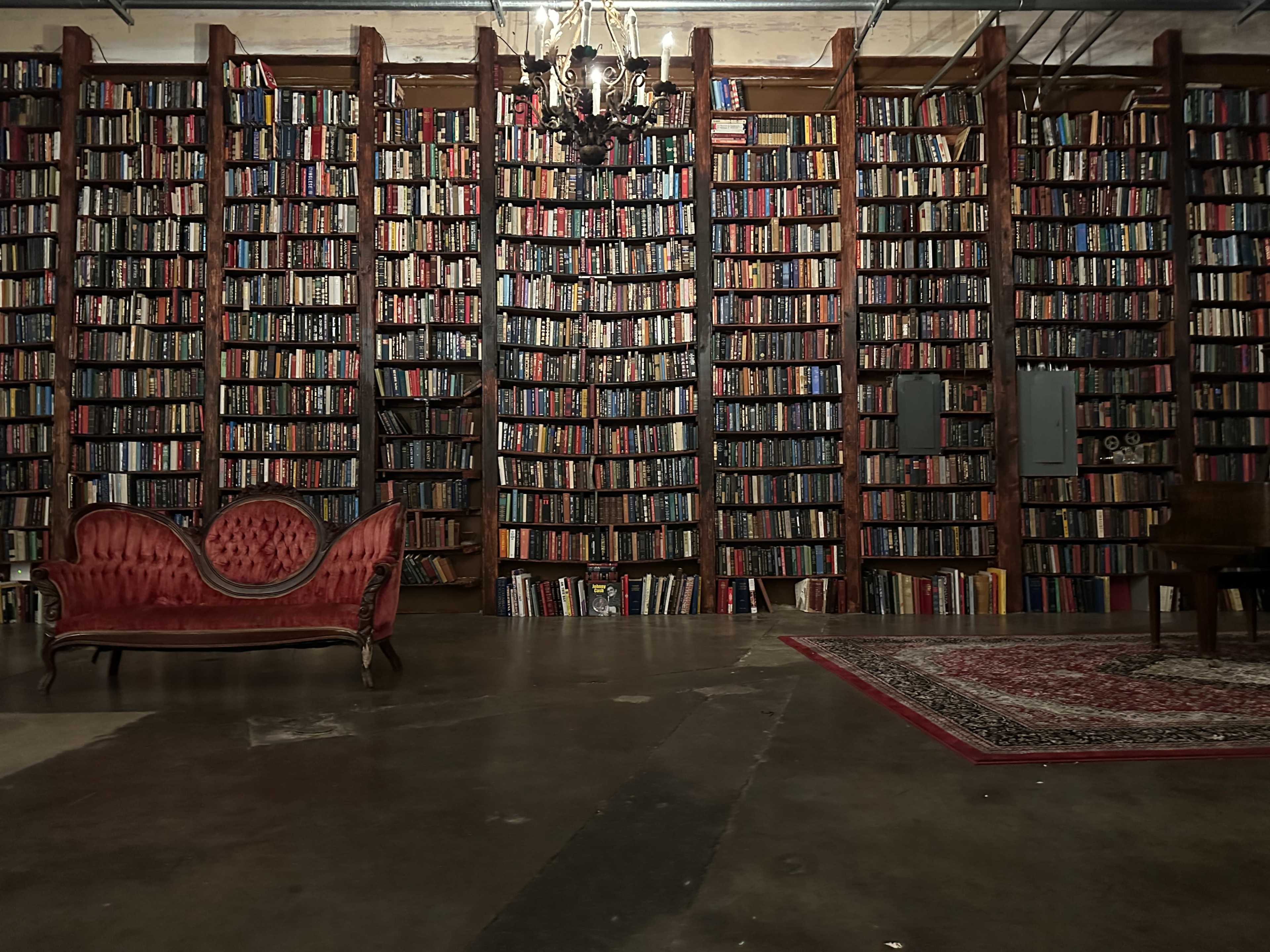 A bookshelf filled with numerous books lines the walls of a dimly lit room, accompanied by a vintage red couch and a small side table.