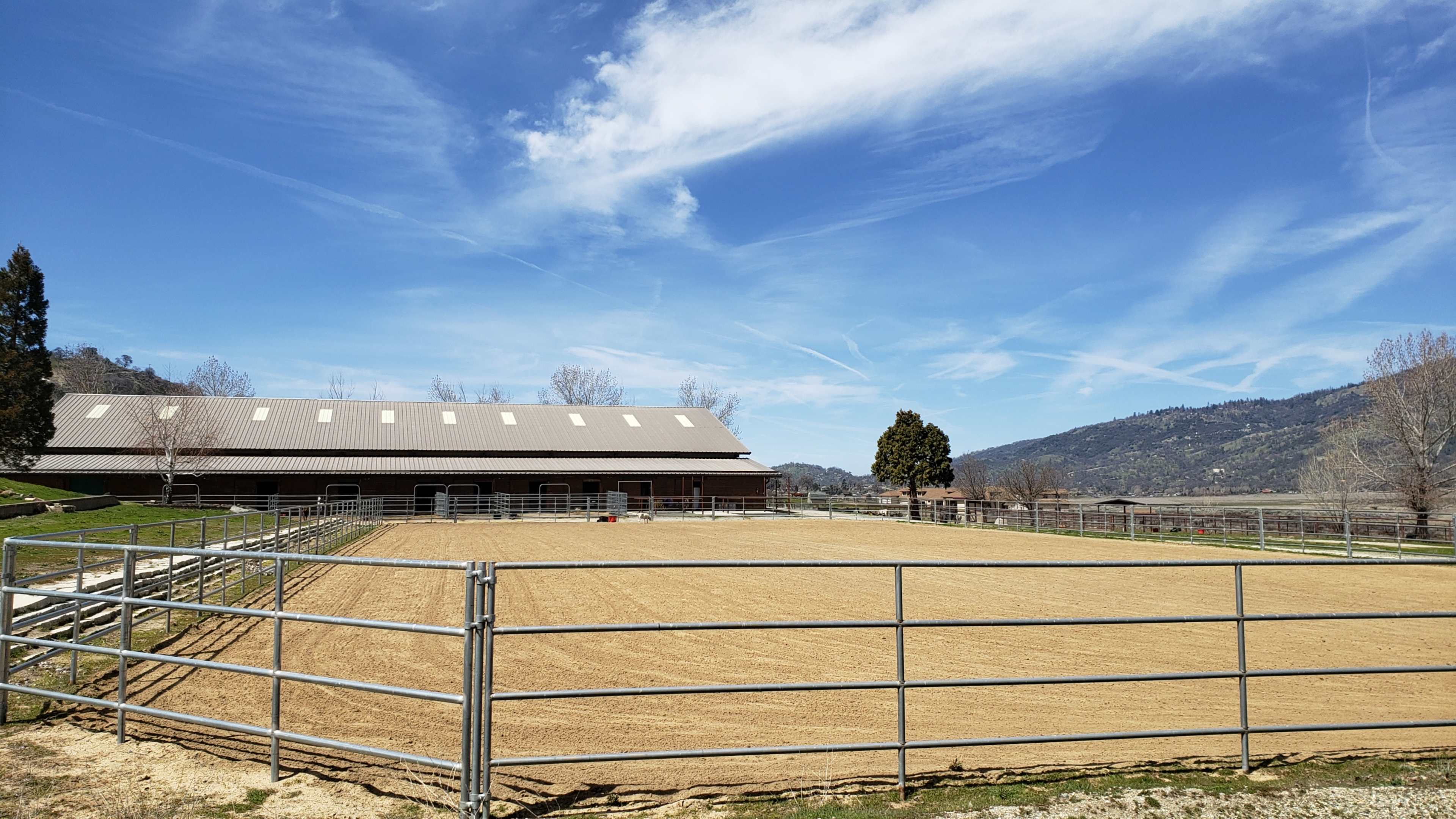 A ranch area with a fenced riding arena, a large barn, and a backdrop of rolling hills under a blue sky.
