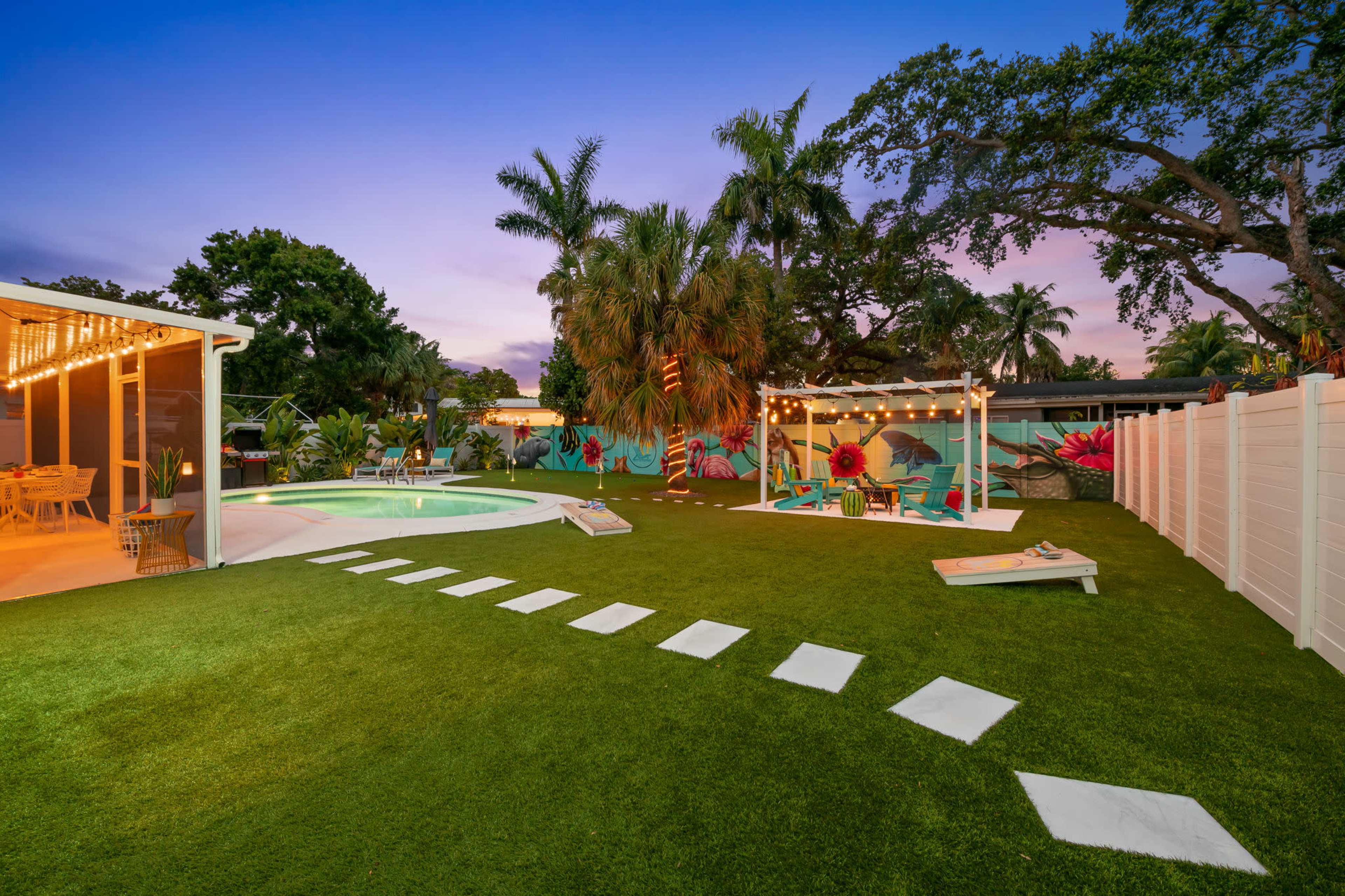 The image depicts a landscaped backyard featuring a swimming pool, a grassy area with stepping stones, and seating arrangements illuminated by decorative string lights.
