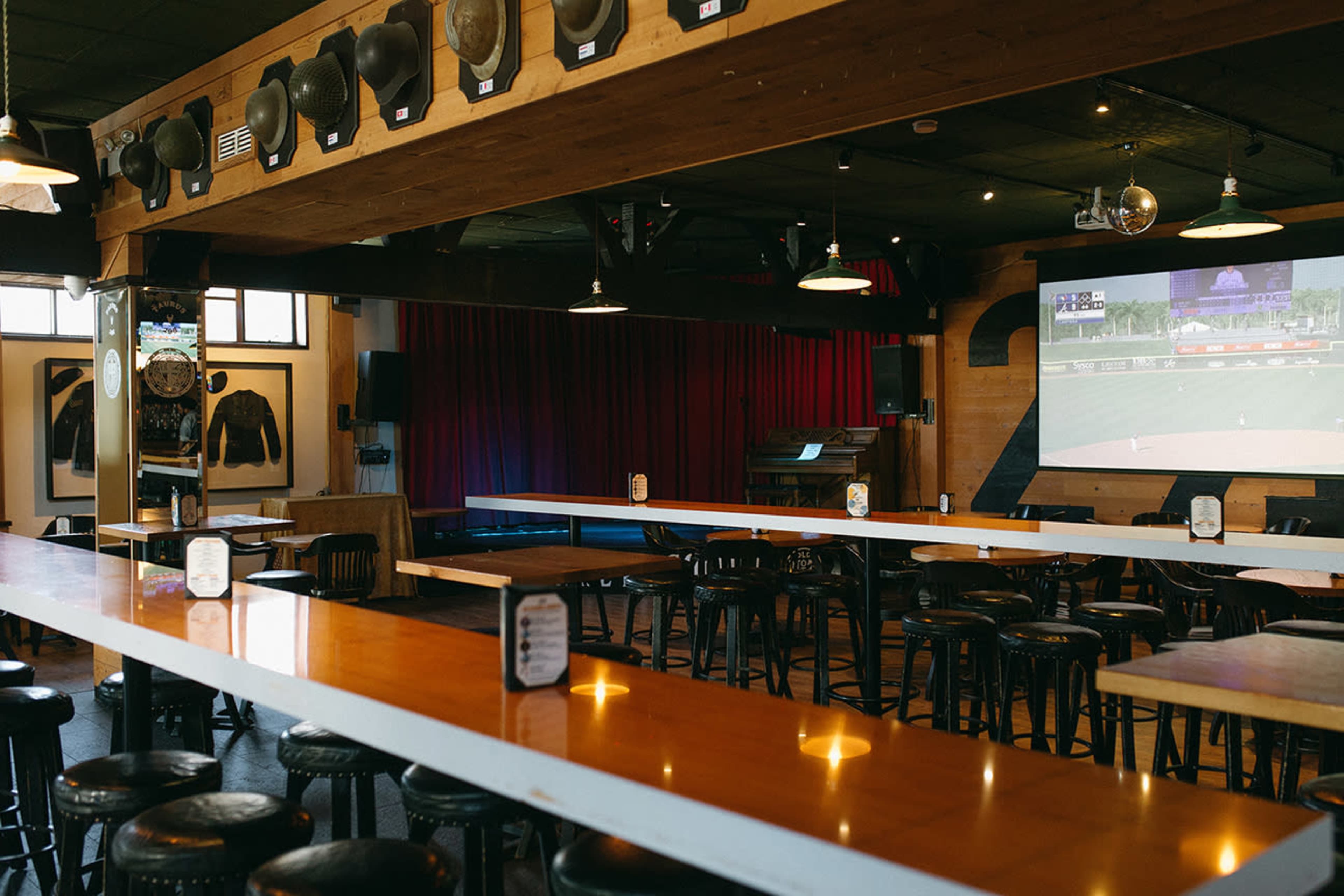 The image shows a spacious bar area with a long wooden counter, black stools, and a large screen displaying a sports game on one wall.