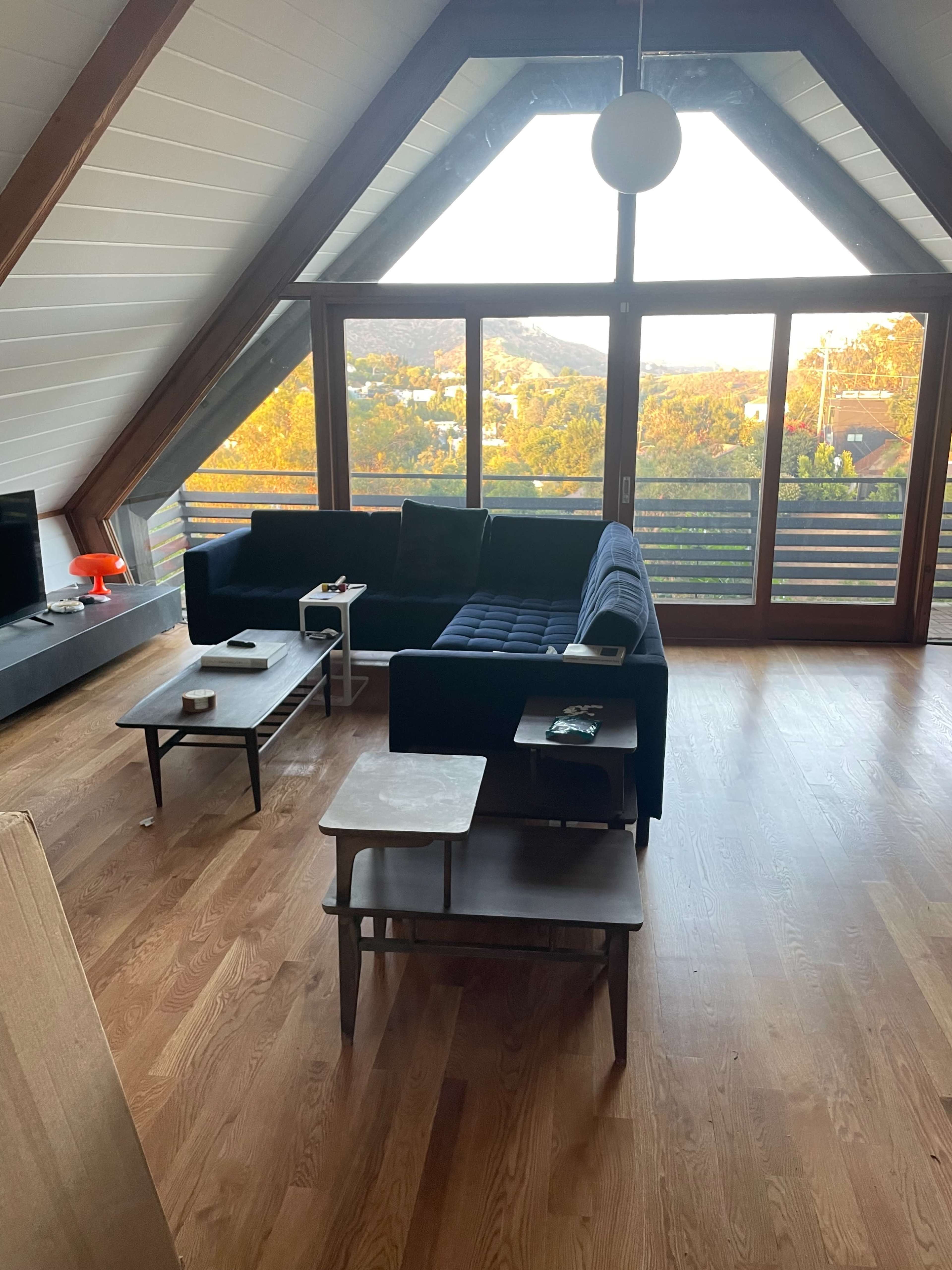 The image shows a modern living room with a blue couch, two wooden coffee tables, and large windows providing a view of the outside landscape.