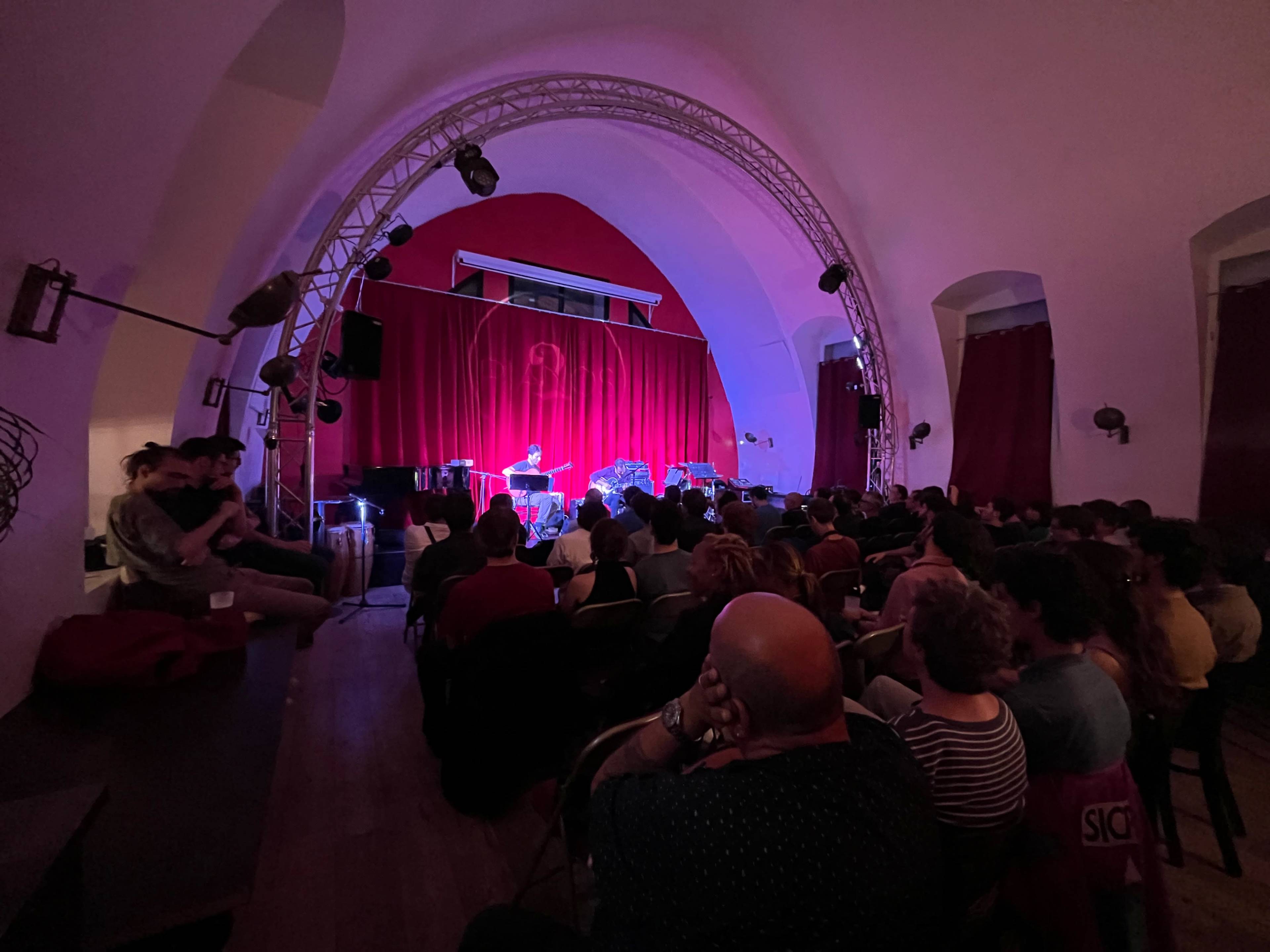 A performer stands on stage in front of a red velvet backdrop, while an audience sits in rows facing them in a dimly lit venue.
