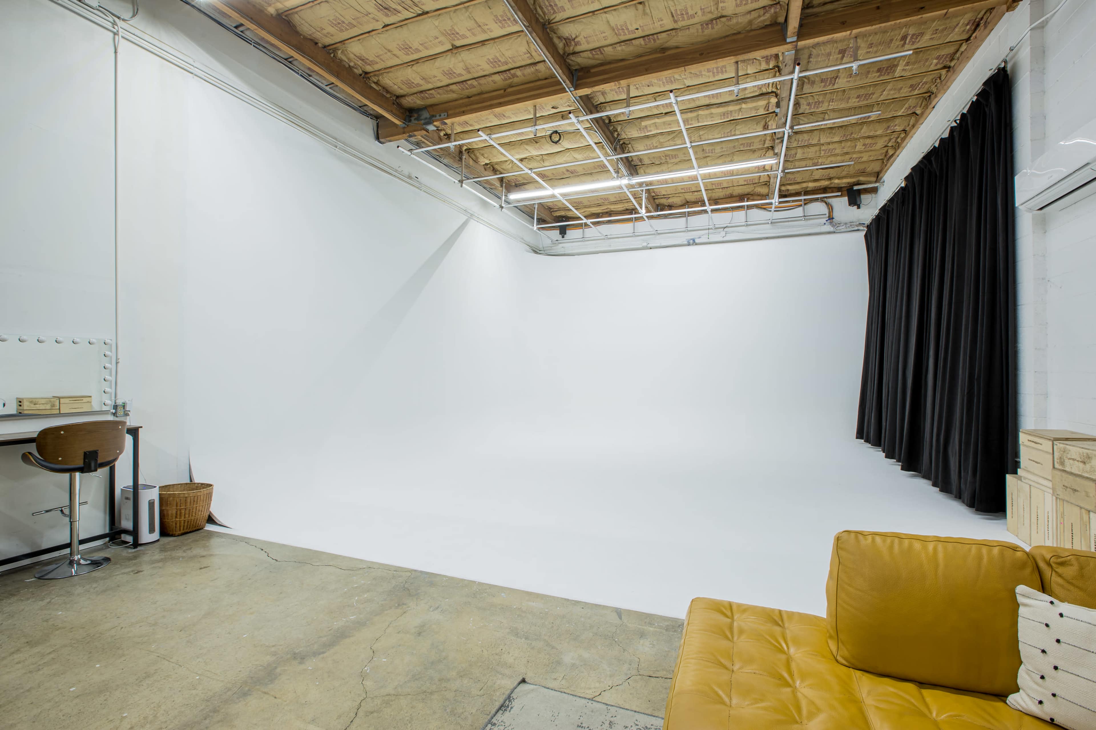 The image shows a spacious photography studio featuring a white backdrop, a yellow couch, a wooden desk with a chair, and a black curtain.