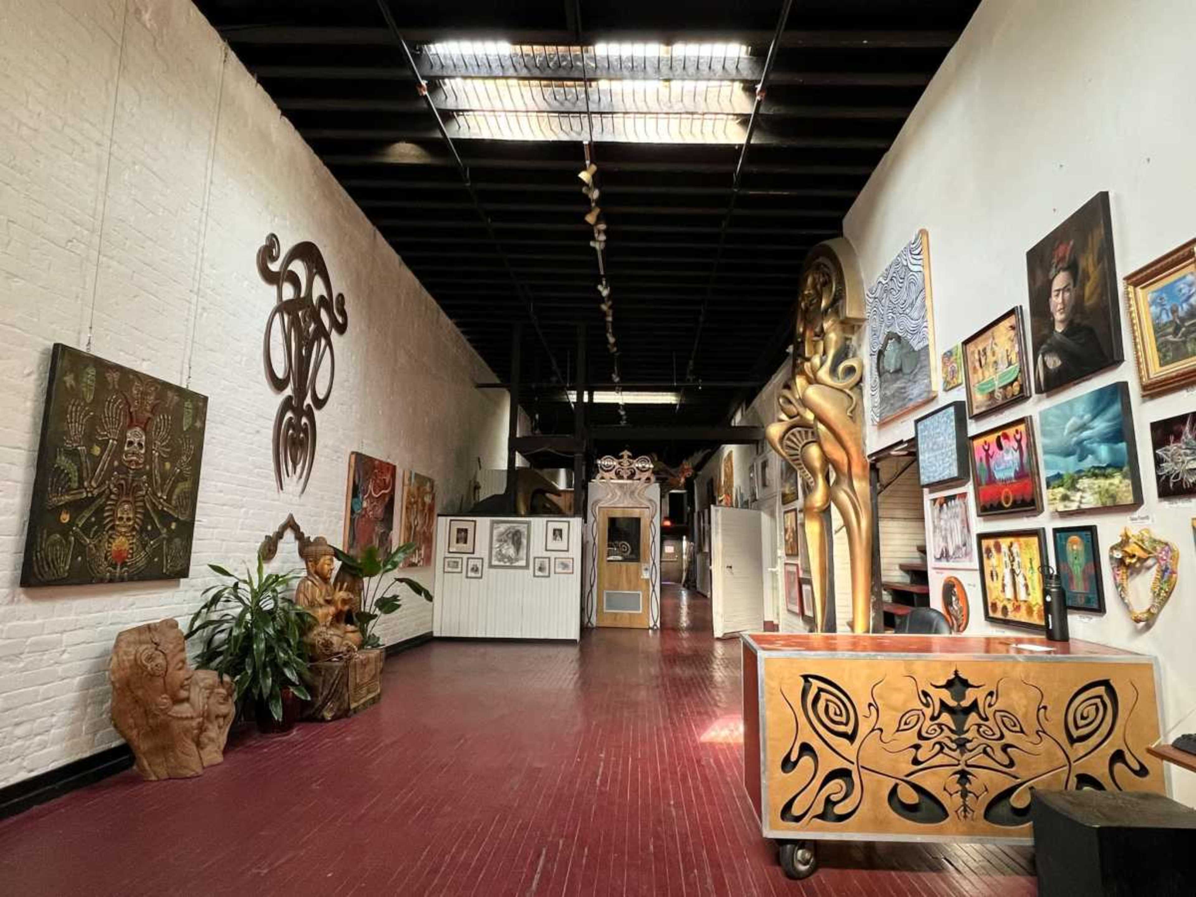 The image shows an art gallery hallway adorned with a variety of paintings, sculptures, and decorative art pieces on the walls and floor.