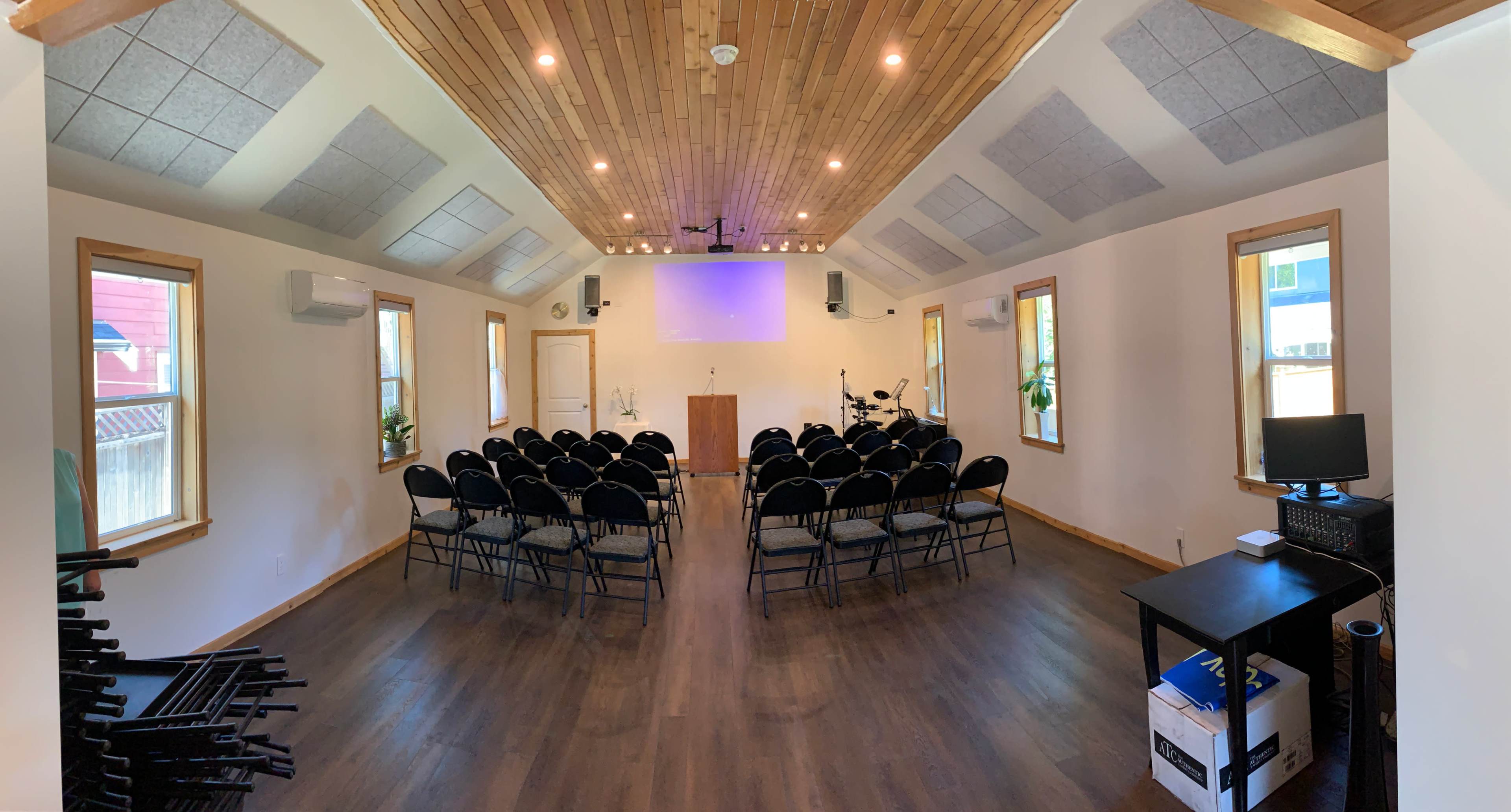 An interior of a small auditorium with rows of black chairs, a podium at the front, and a sound system setup.