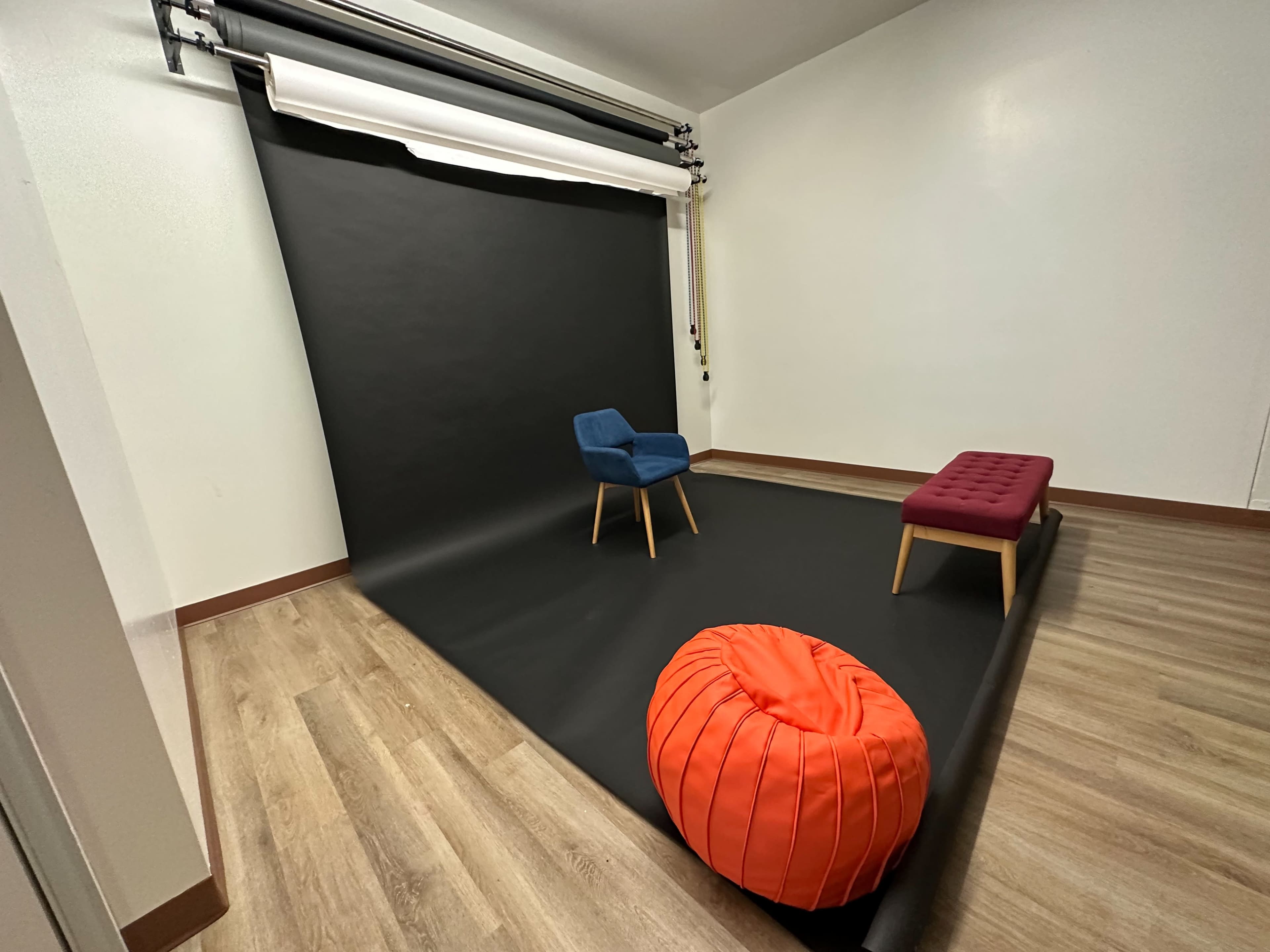 A minimalist photography studio featuring a black backdrop, a blue chair, a red ottoman, and an orange beanbag on wooden flooring.