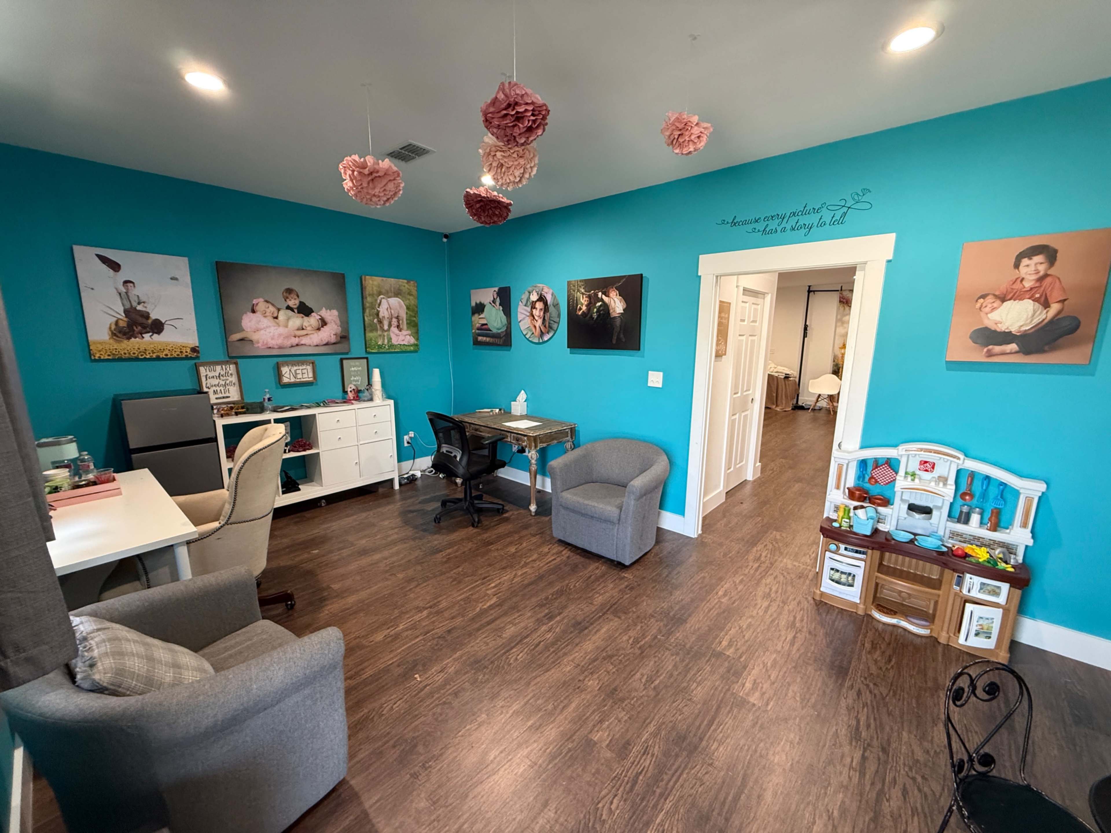 The image shows a brightly colored room with teal walls, featuring two armchairs, a desk with a computer, and various framed photographs on display.