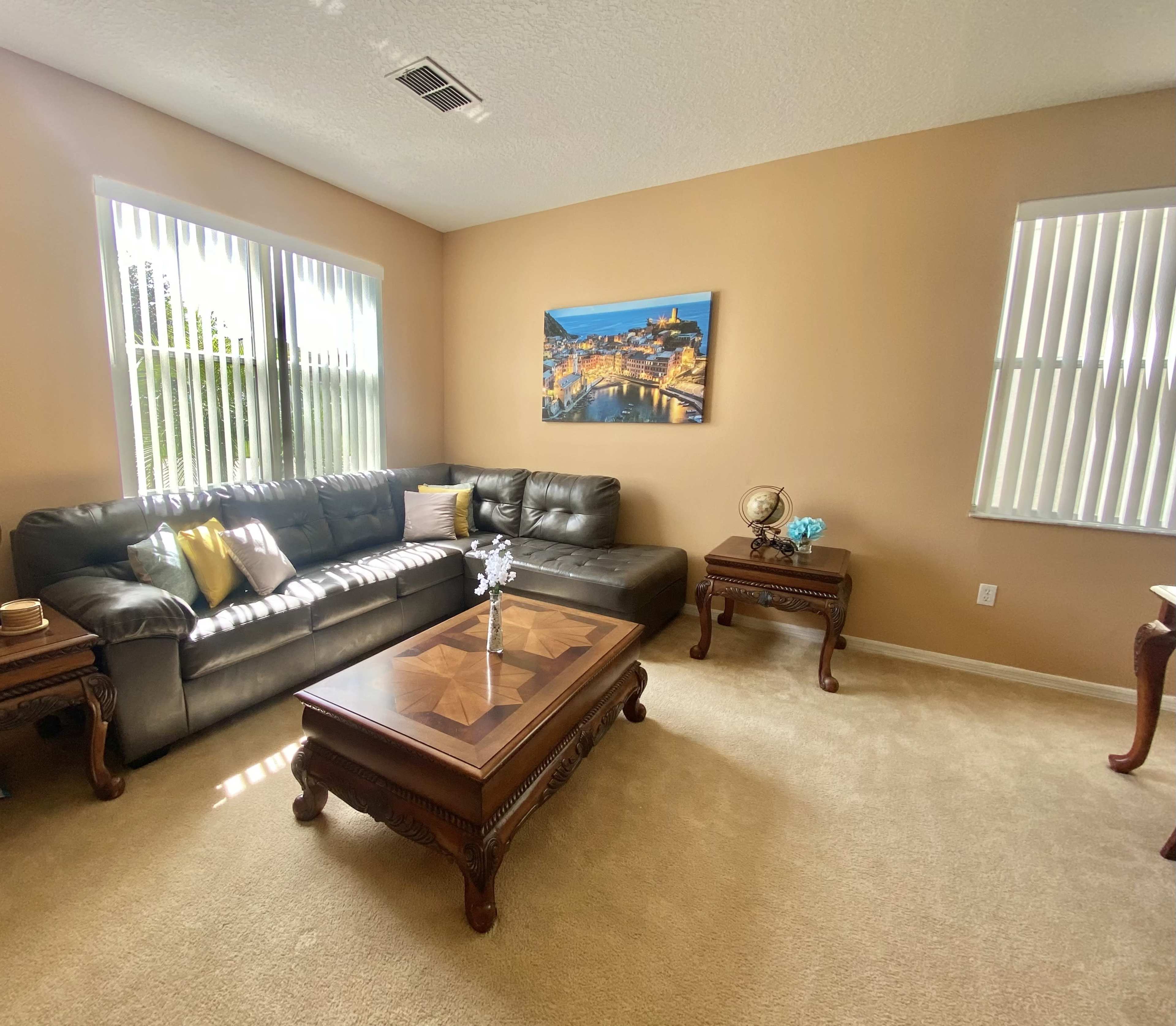 The room features a modern dark leather sectional sofa, a wooden coffee table with intricate details, and a landscape painting on the wall, with light filtering through the window blinds.