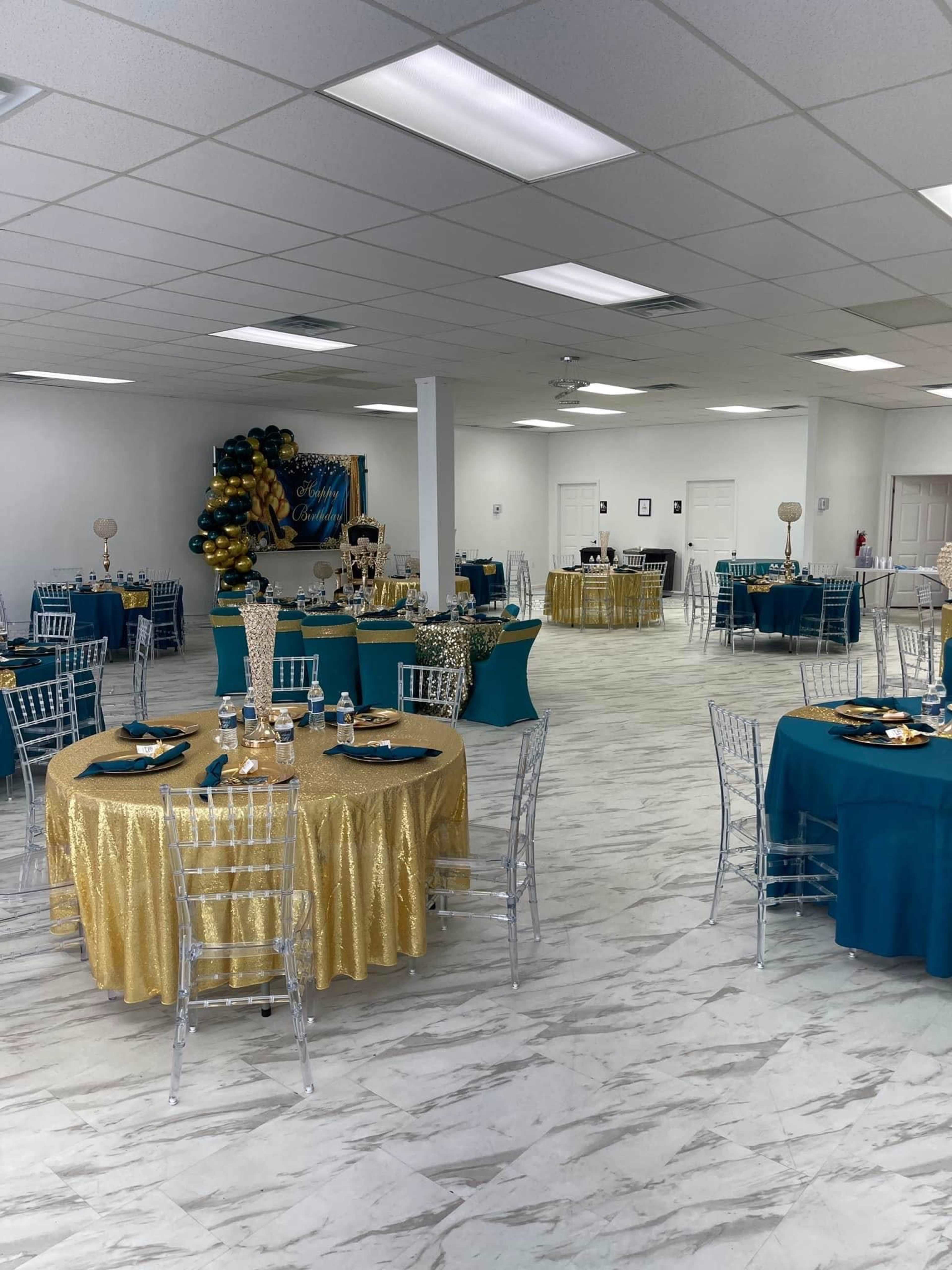A spacious event hall with round tables dressed in gold and teal tablecloths, surrounded by decorative balloons and a backdrop.