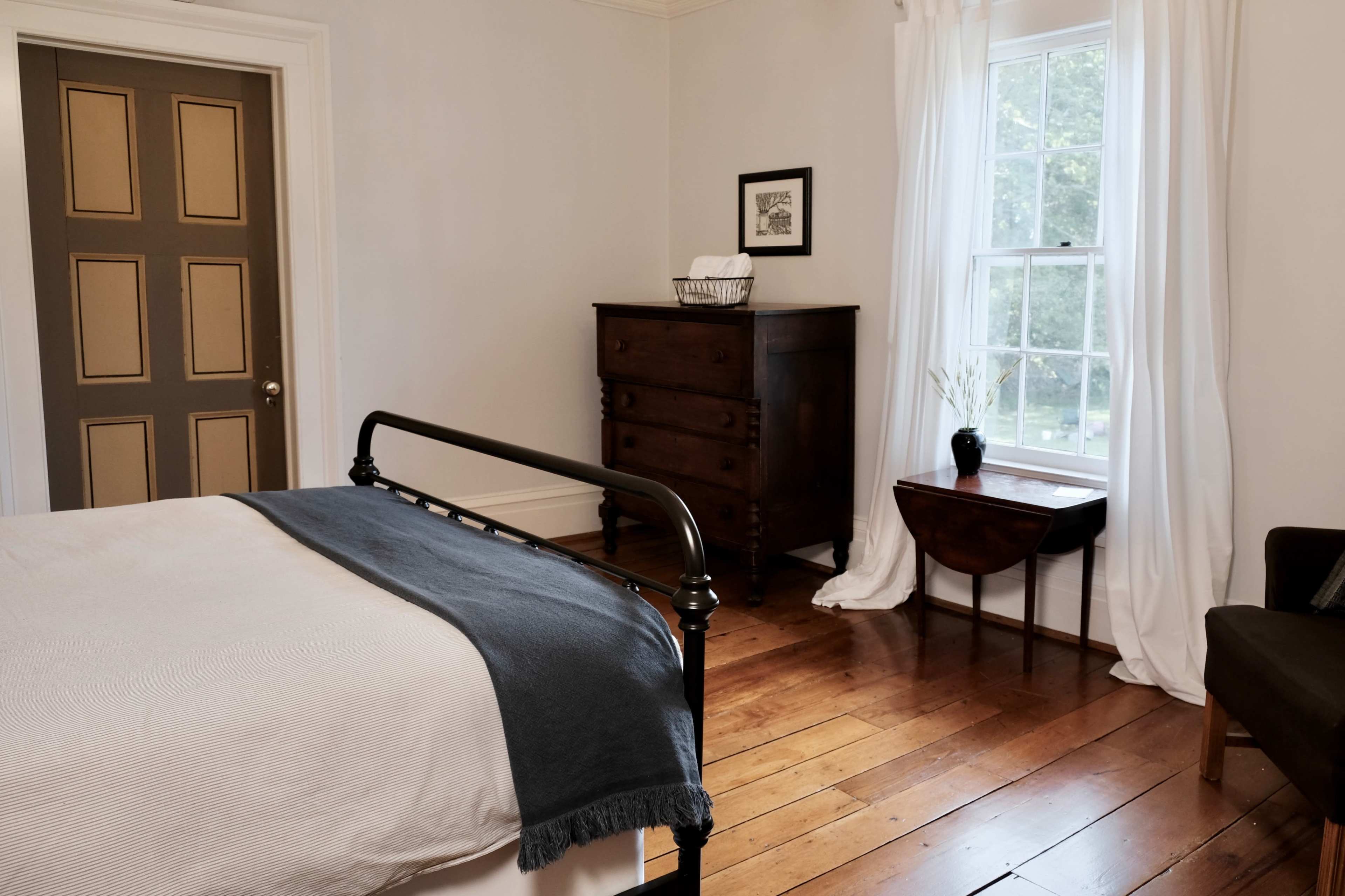 A simple bedroom with a bed, a dresser, a small table, and a large window letting in natural light.