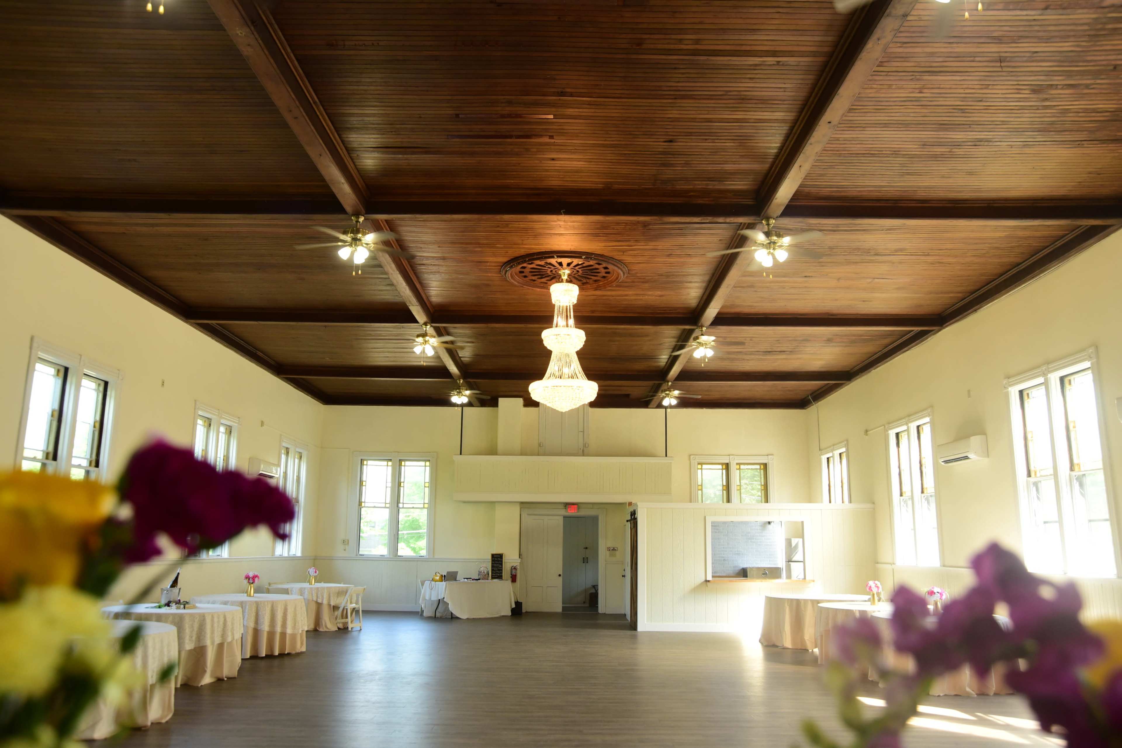 The image shows a spacious event hall with a wooden ceiling, a chandelier, and several tables arranged around the perimeter.
