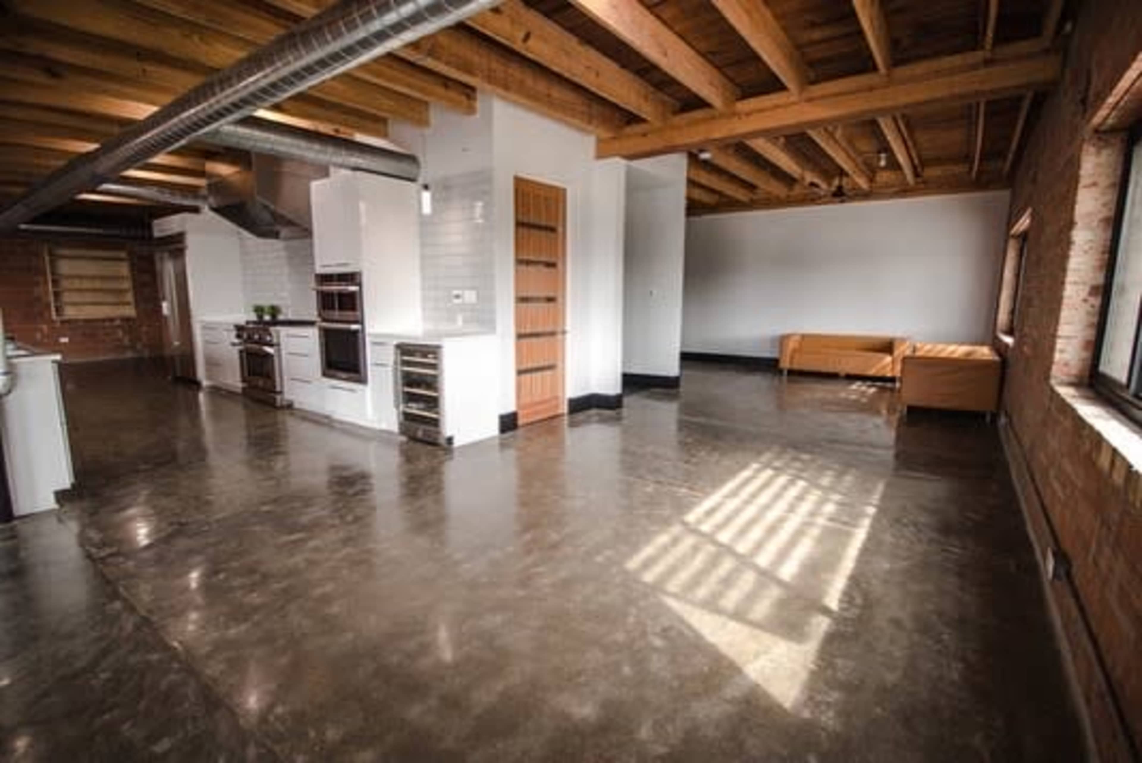 The image shows a spacious industrial-style kitchen and living area with polished concrete floors, exposed wooden beams, and minimalistic furnishings.