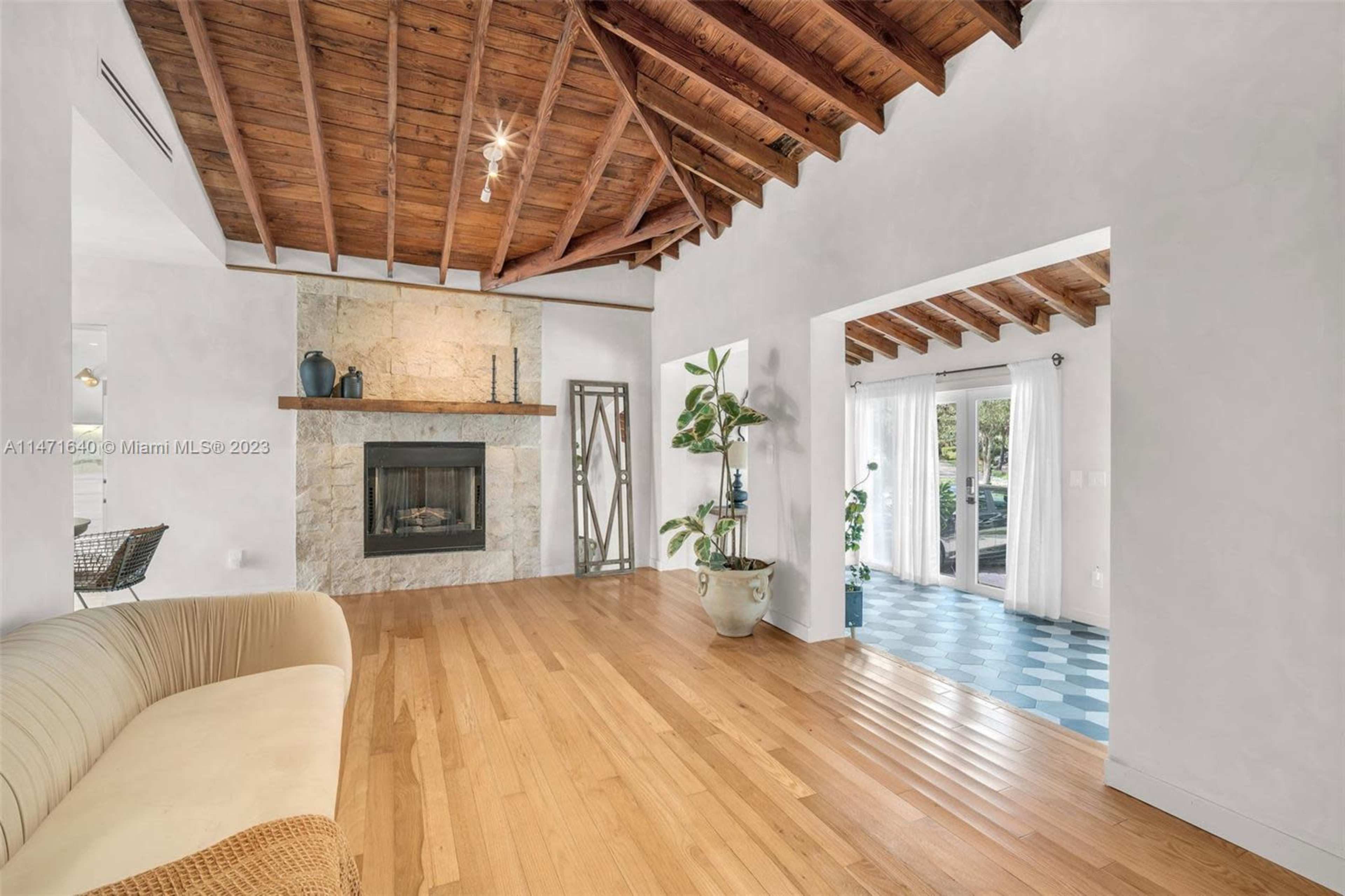 The image shows a living room with a stone fireplace, wooden ceiling beams, and a large window that opens to an outdoor area.