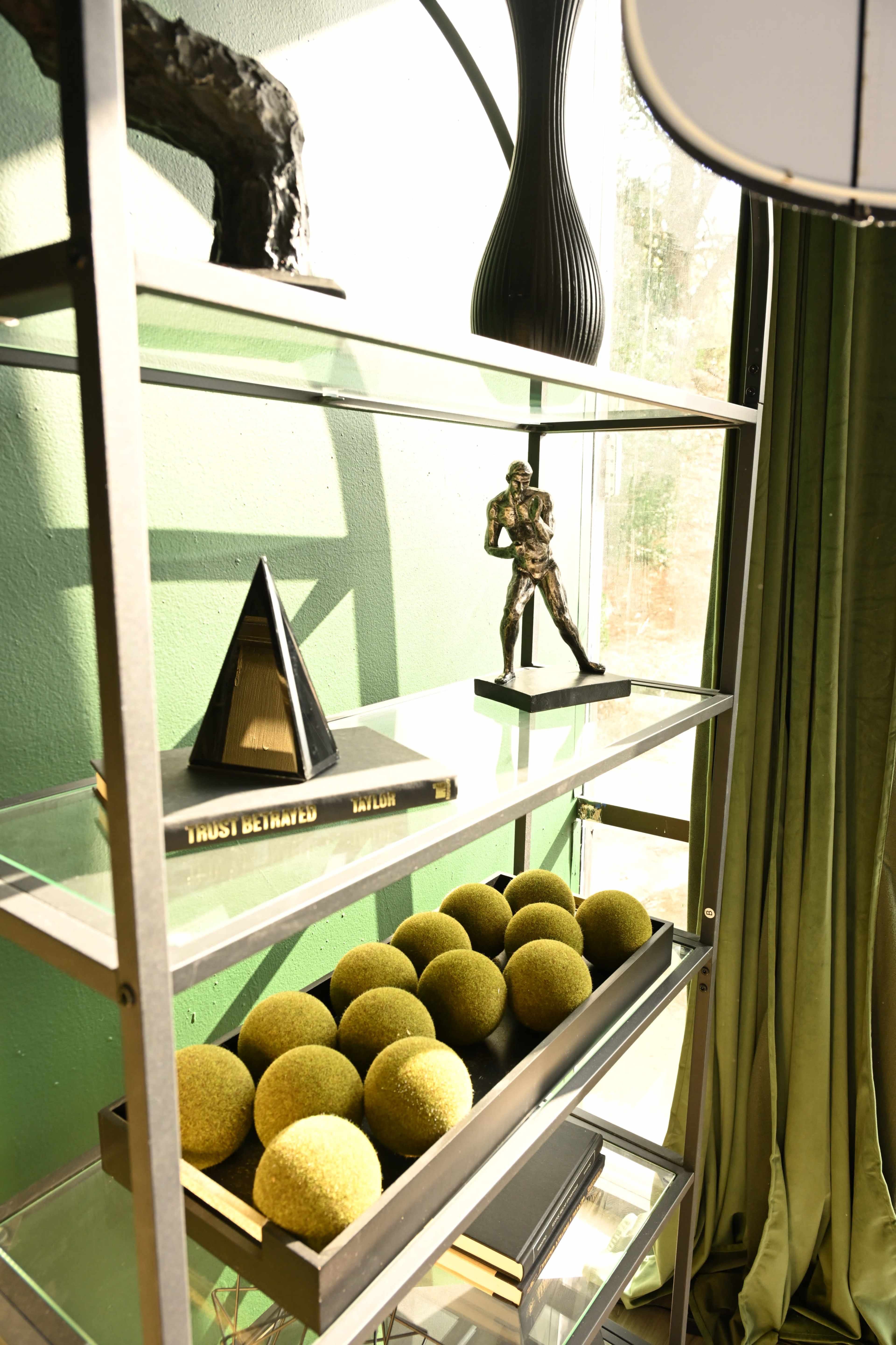 A metal shelf displays a triangular sculpture, a small figurine, a book, and a collection of green spherical objects, set against a green wall and curtains.