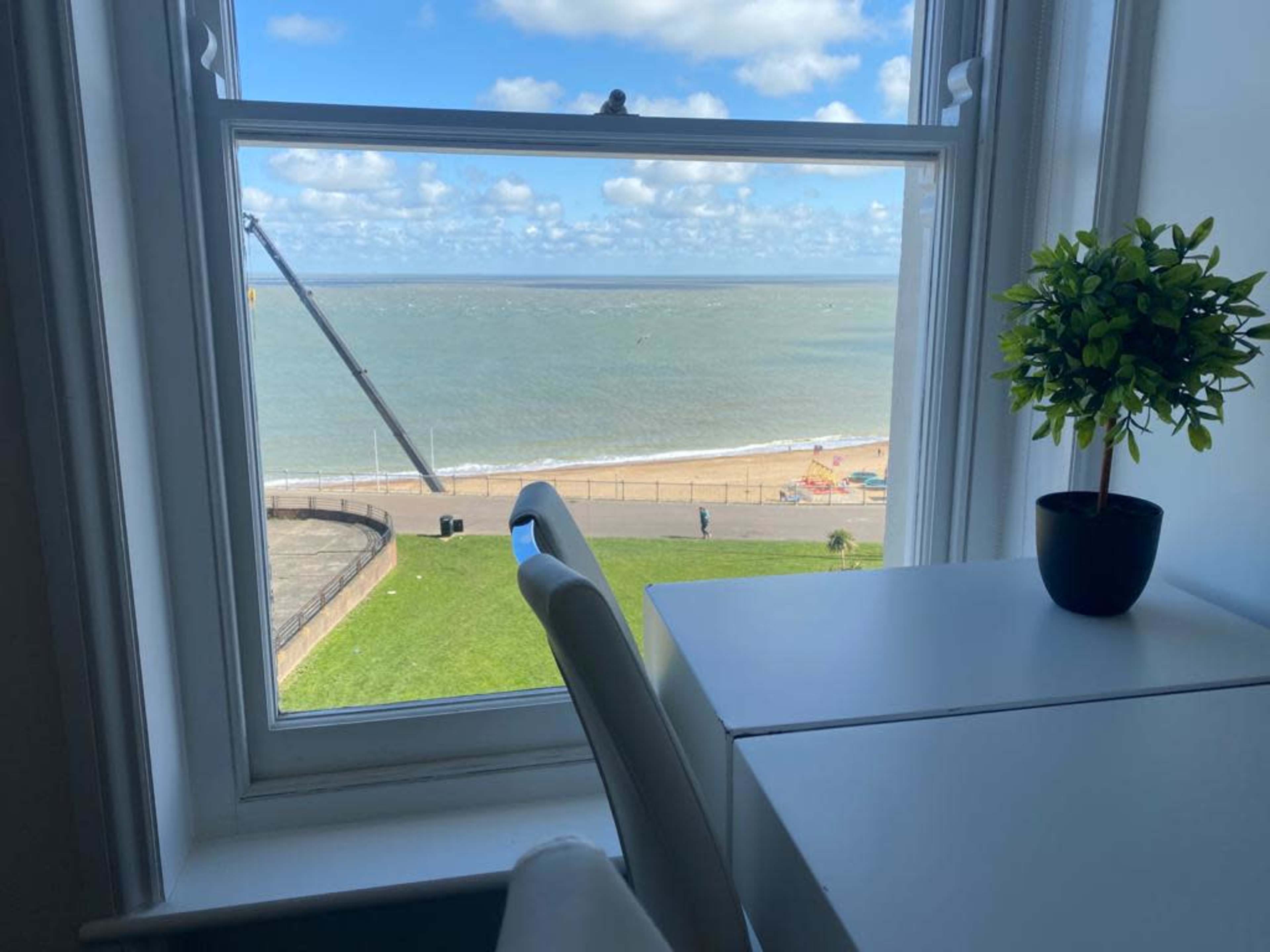 The scene shows a view from a window overlooking a beach with a sea and clouds in the sky, accompanied by a small plant on a white desk.