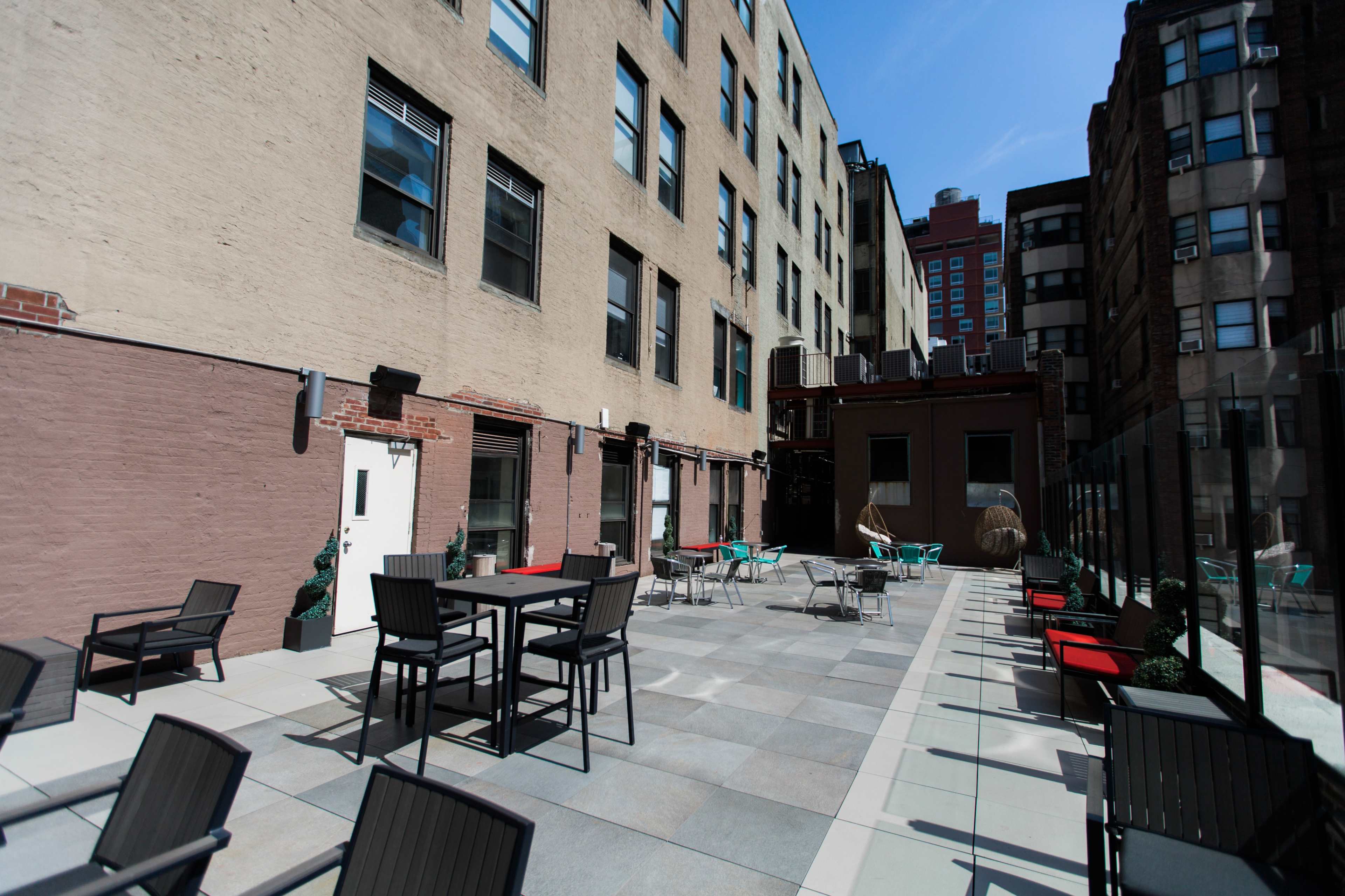 The image shows a rooftop patio area with tables and chairs, surrounded by brick and concrete buildings under a clear blue sky.
