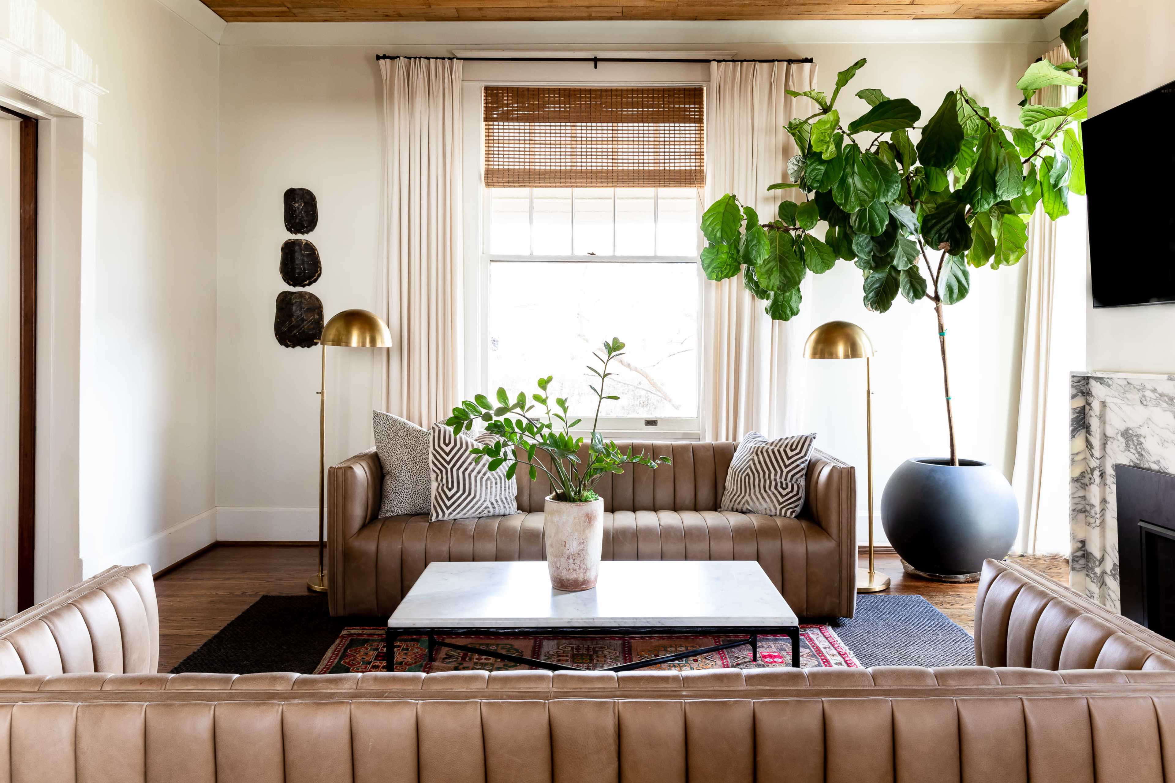 The image shows a modern living room with two brown leather sofas, a marble coffee table, potted plants, and a large window draped with sheer curtains.