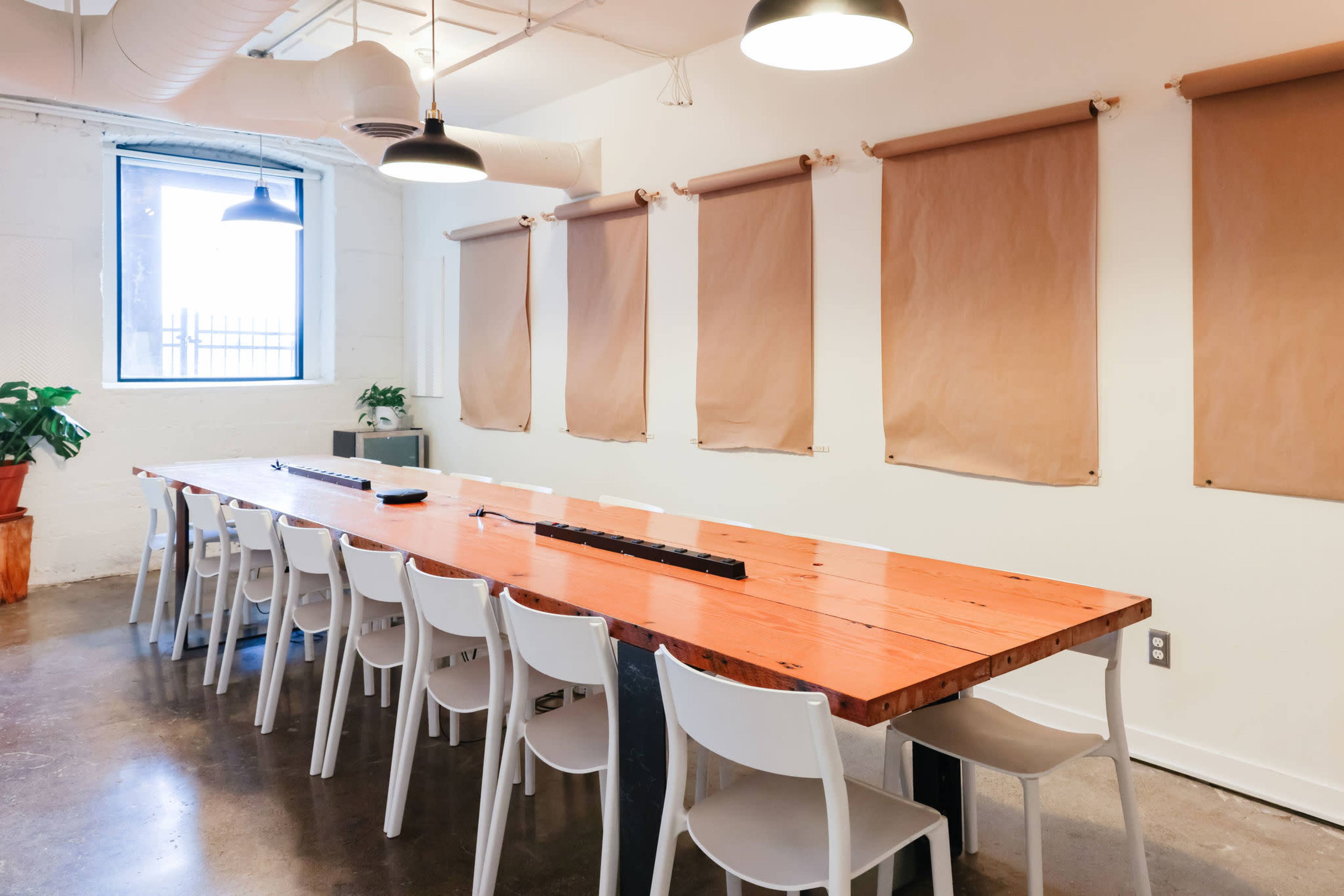 A large, wooden table surrounded by white chairs is positioned in a well-lit meeting room with brown roller shades and a potted plant in the corner.
