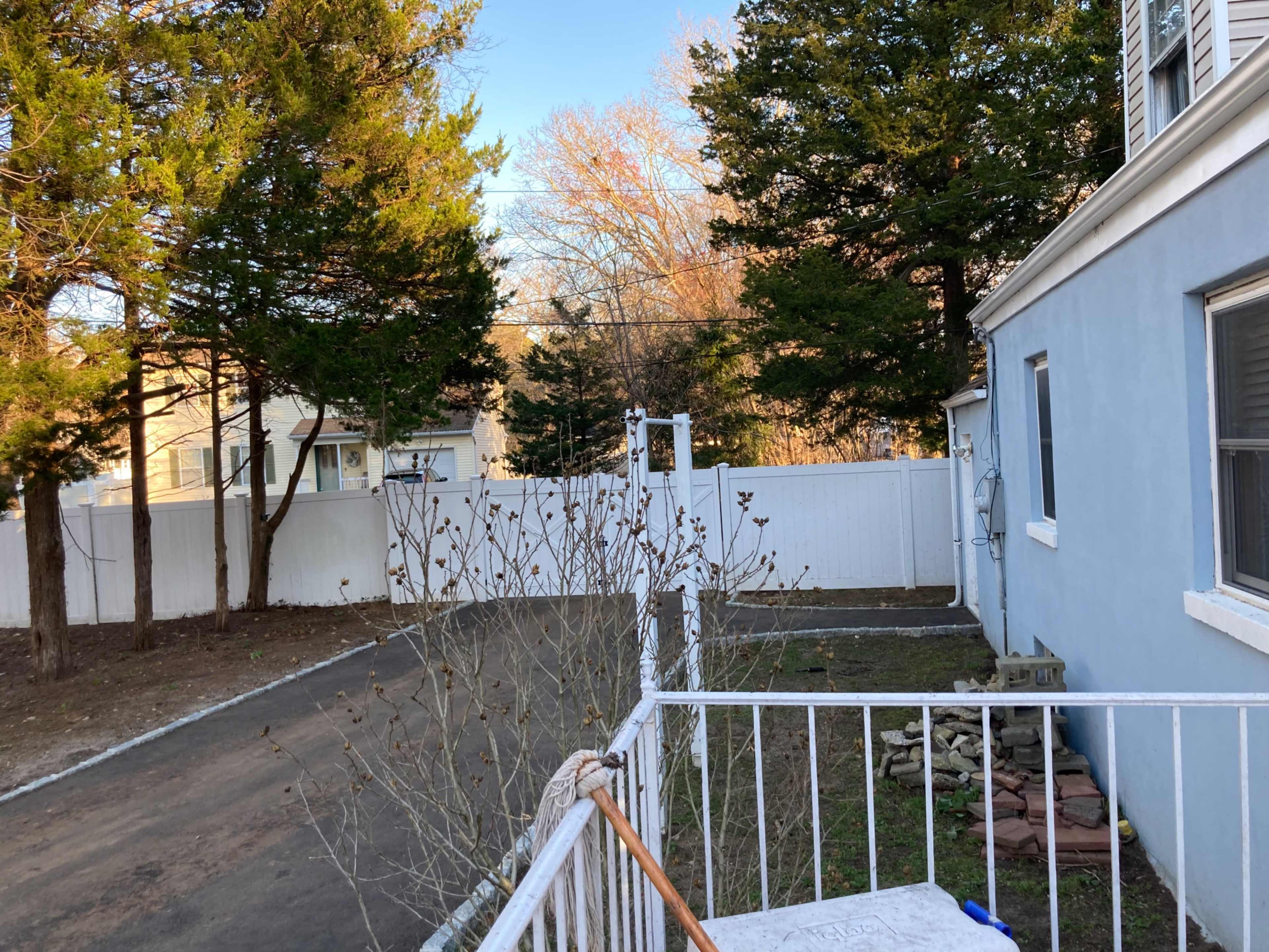 A gravel driveway beside a blue house, bordered by a white fence and trees in the background.