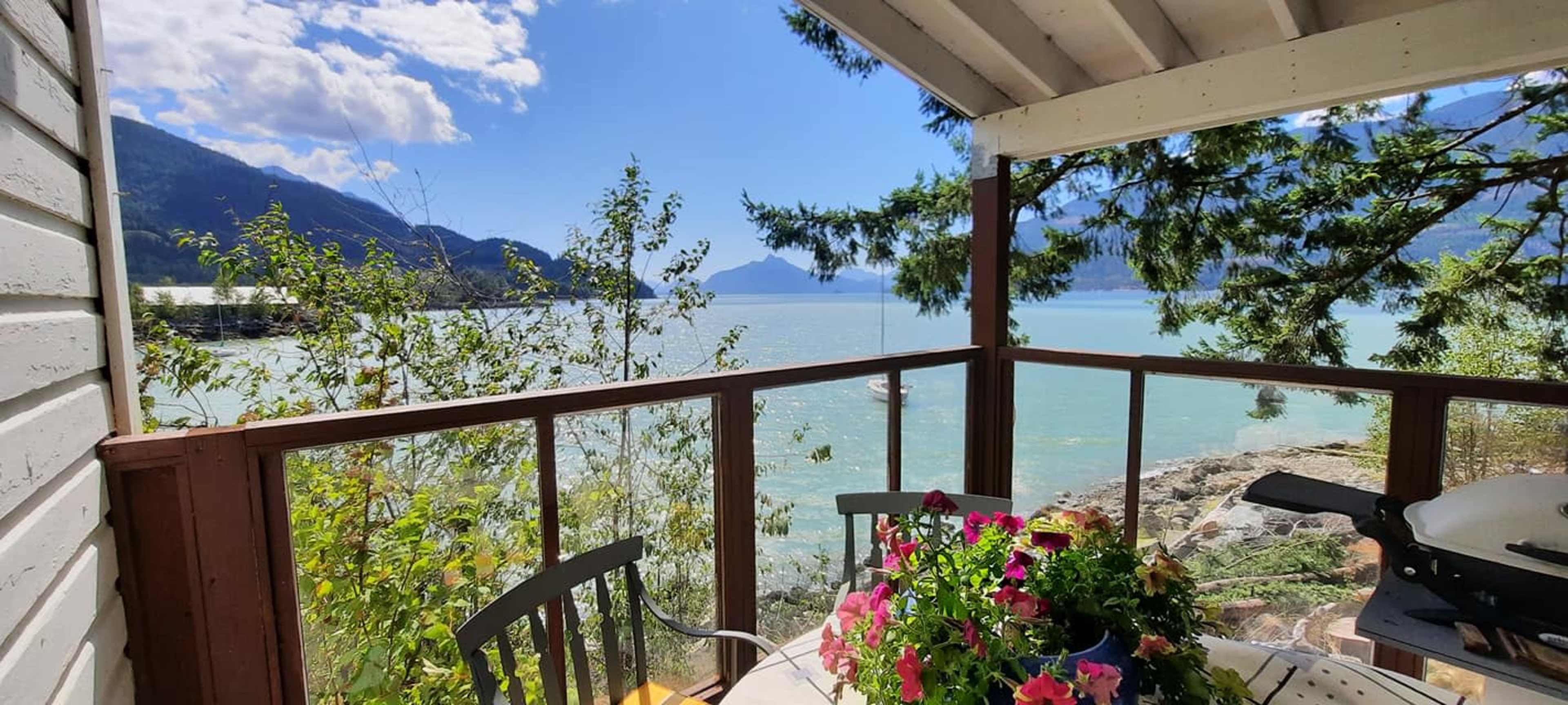 A wooden balcony overlooking a calm body of water surrounded by mountains and trees, with a table adorned with flowering plants.