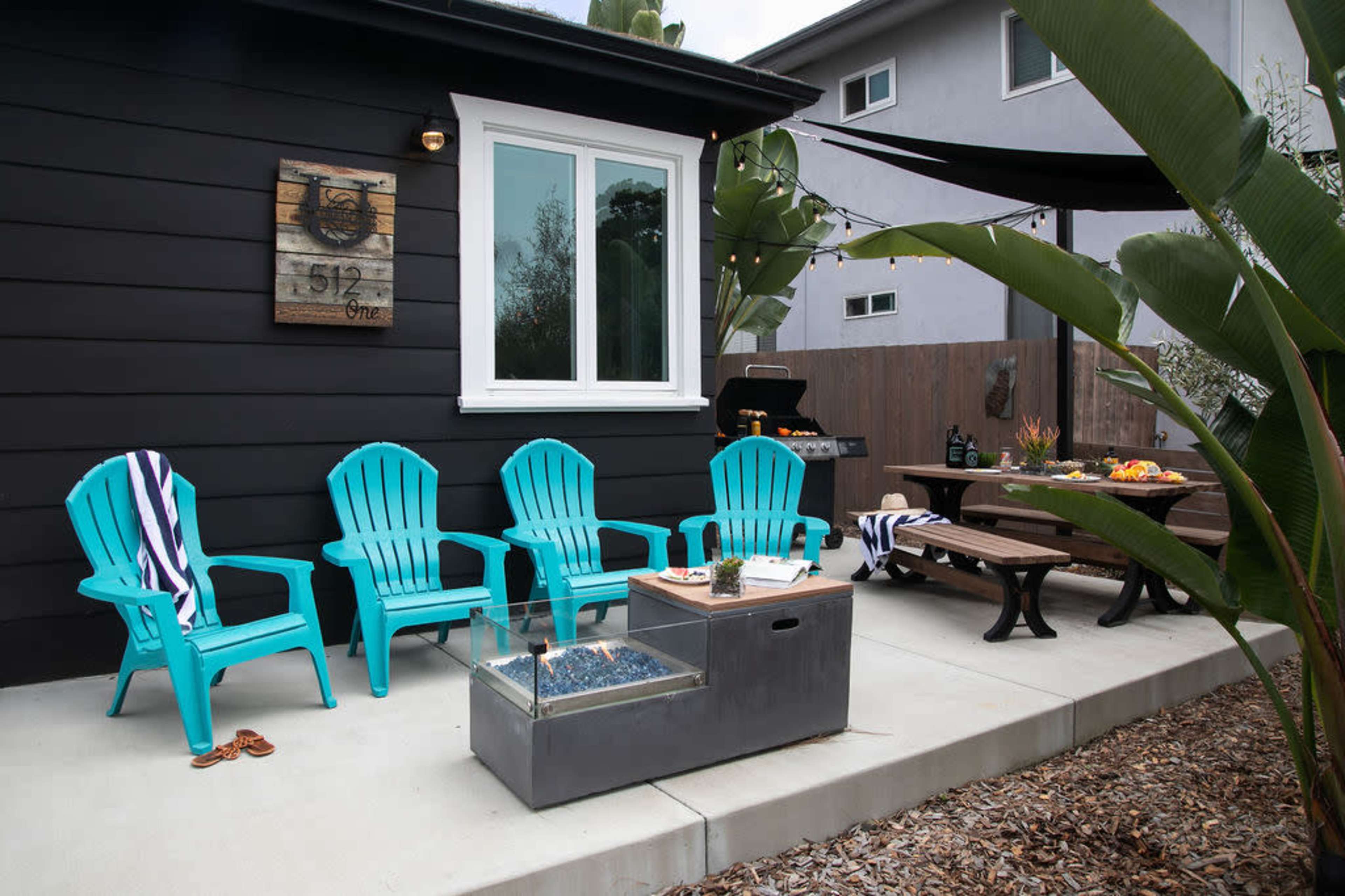 The image shows a outdoor patio area featuring four teal adirondack chairs, a dining table, a grill, and a fire pit, set among plants and a black-shingled house.