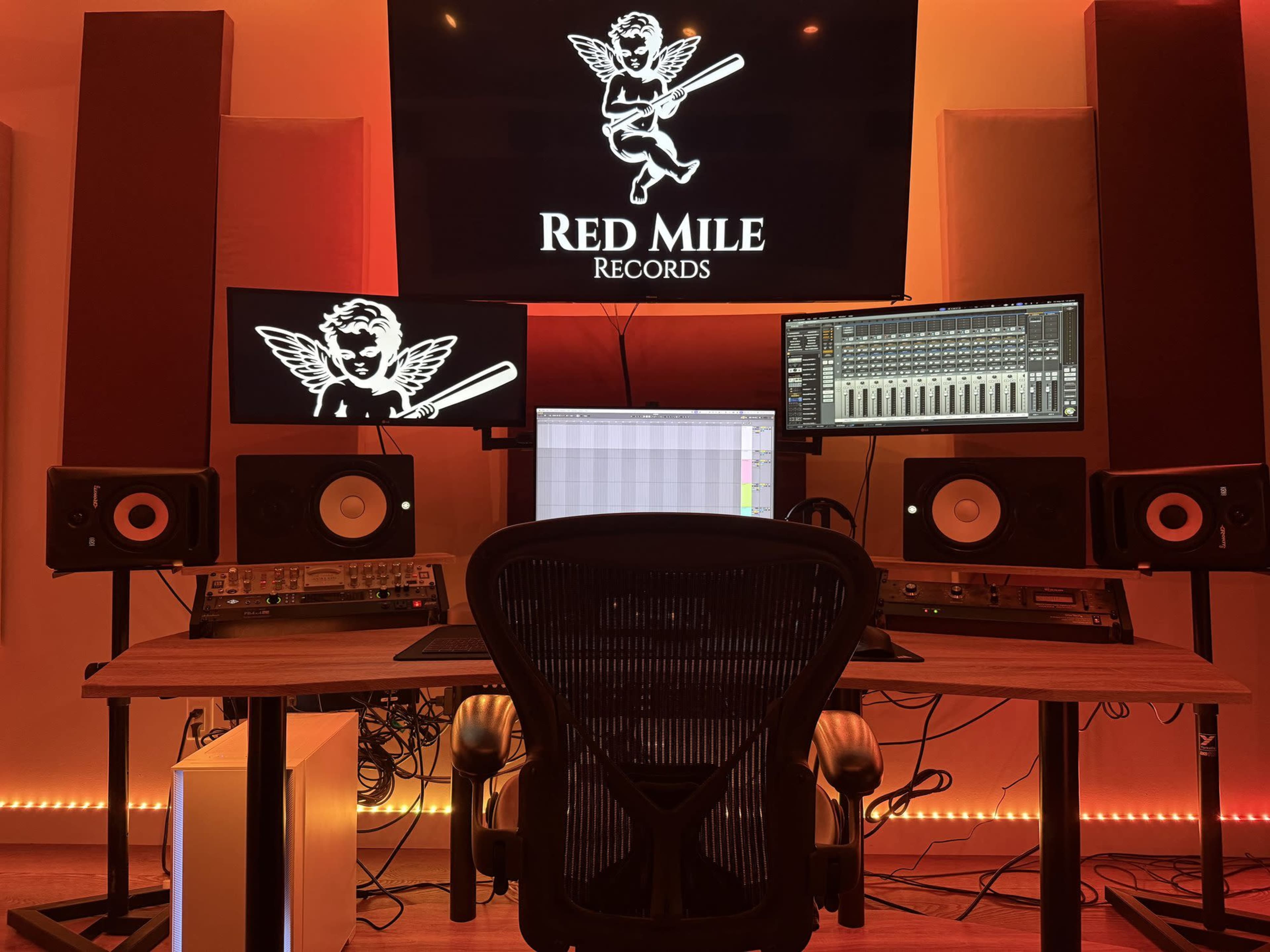 The image shows a music recording studio with a large screen displaying the "Red Mile Records" logo, two monitors, speakers, and an ergonomic chair at the center of a wooden desk.