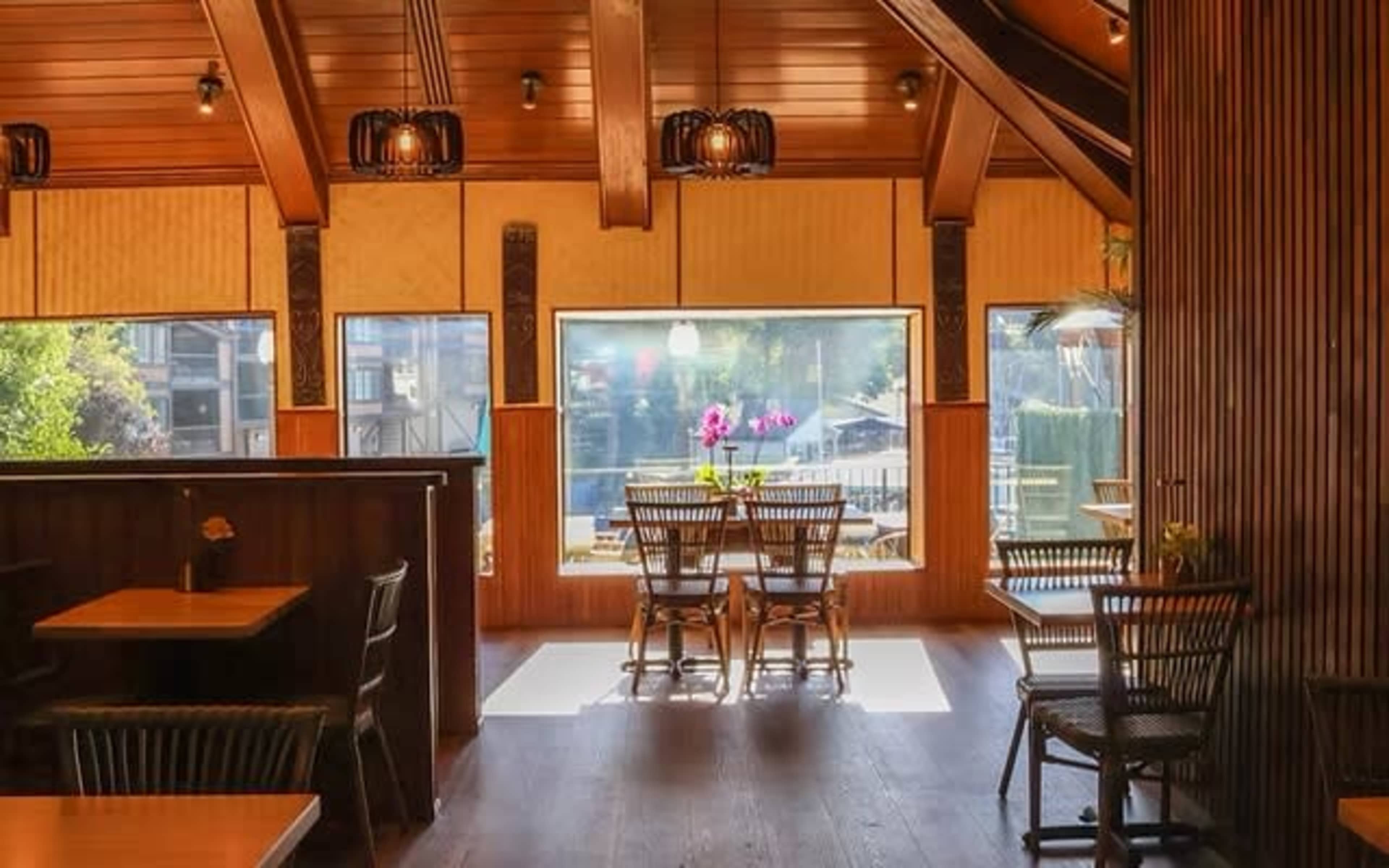 The image shows a wooden interior of a restaurant with large windows overlooking a patio area, featuring tables and chairs arranged for dining.