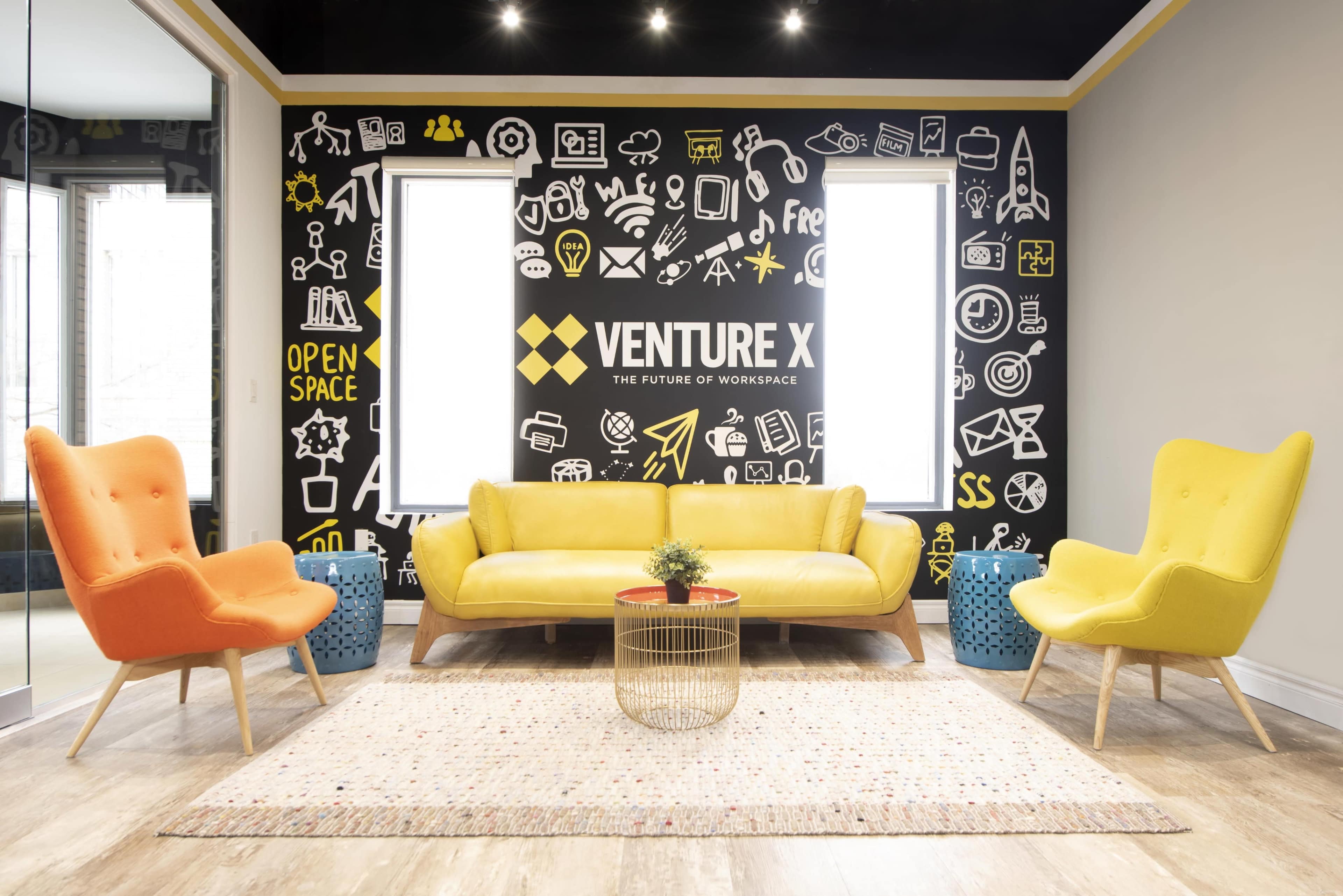 A modern office lounge area with a yellow sofa, orange and yellow armchairs, a round coffee table, and a black wall adorned with various icons related to work and creativity.