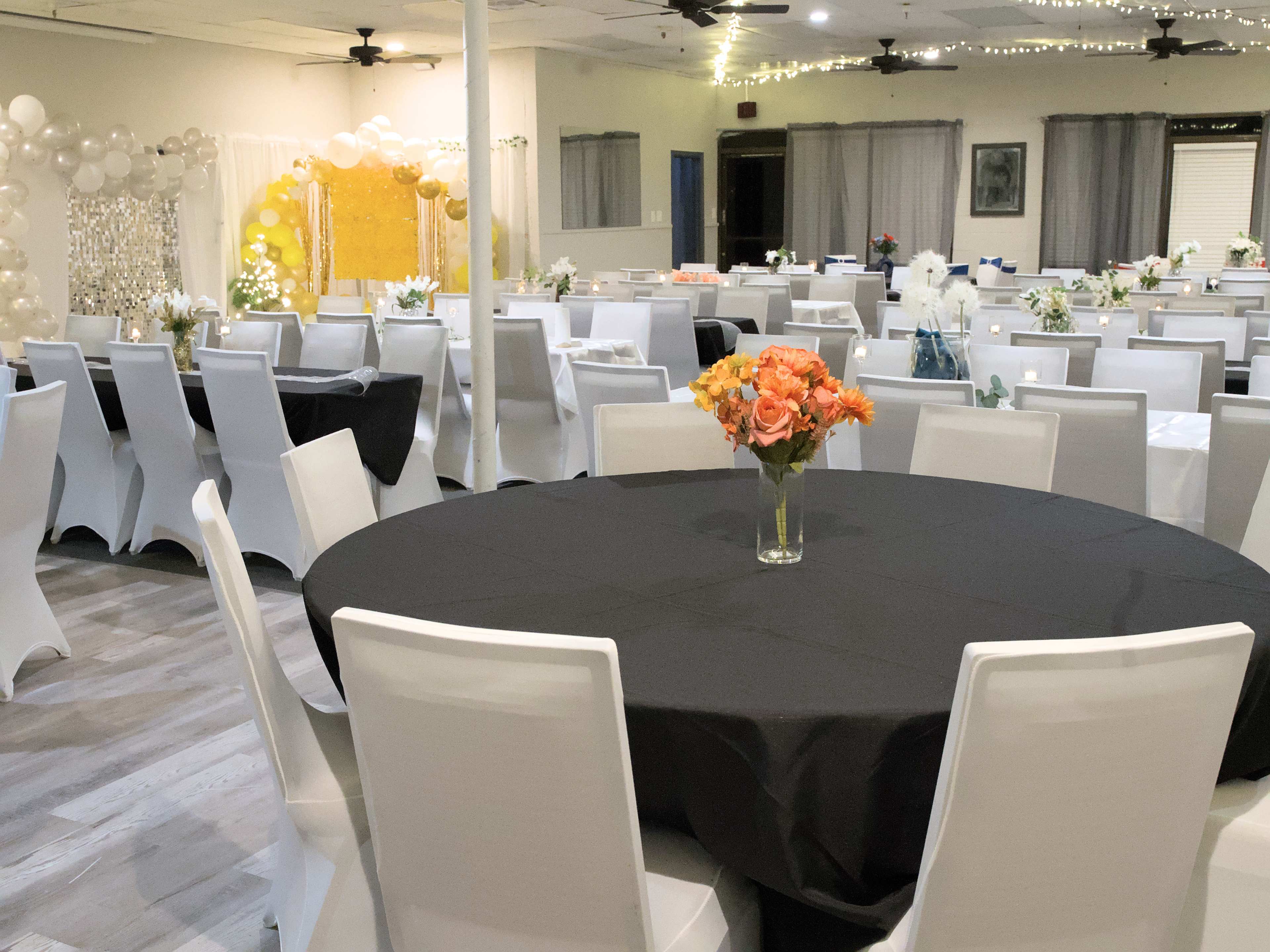 A decorated event space with round tables covered in black and white tablecloths, floral centerpieces, and a backdrop adorned with balloons.