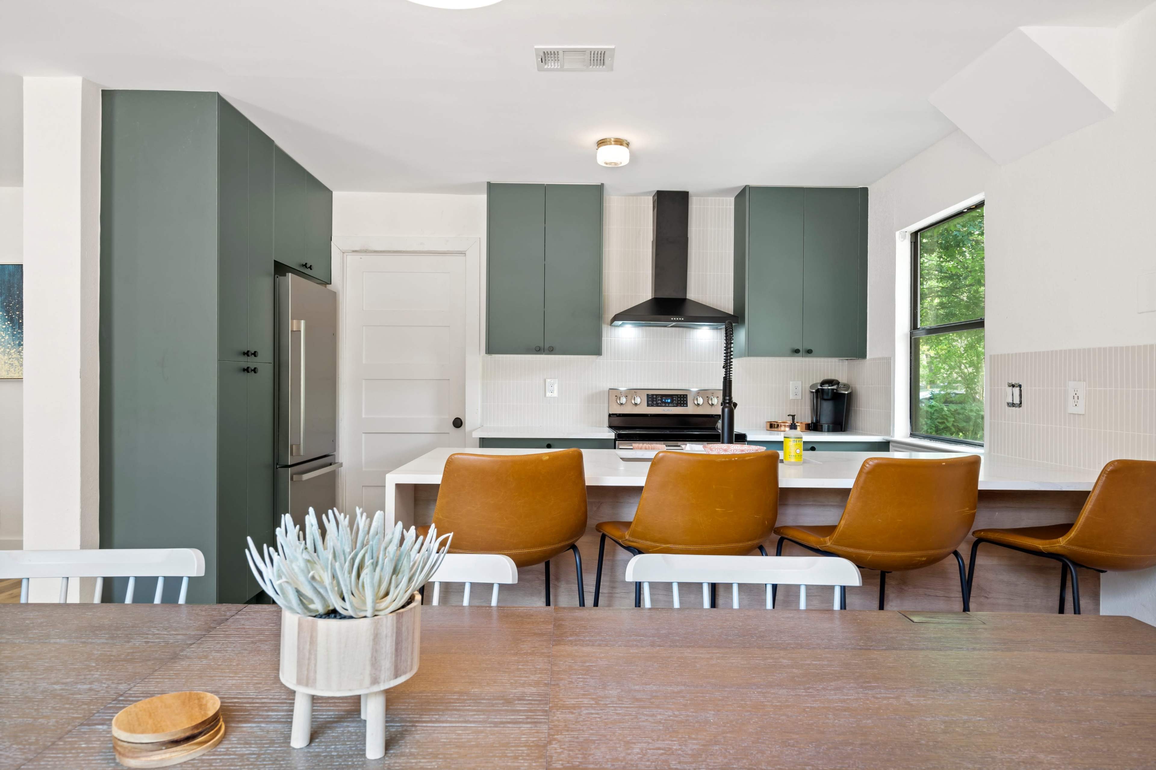 A modern kitchen features green cabinetry, a gray countertop, and a dining table with a small succulent centerpiece.