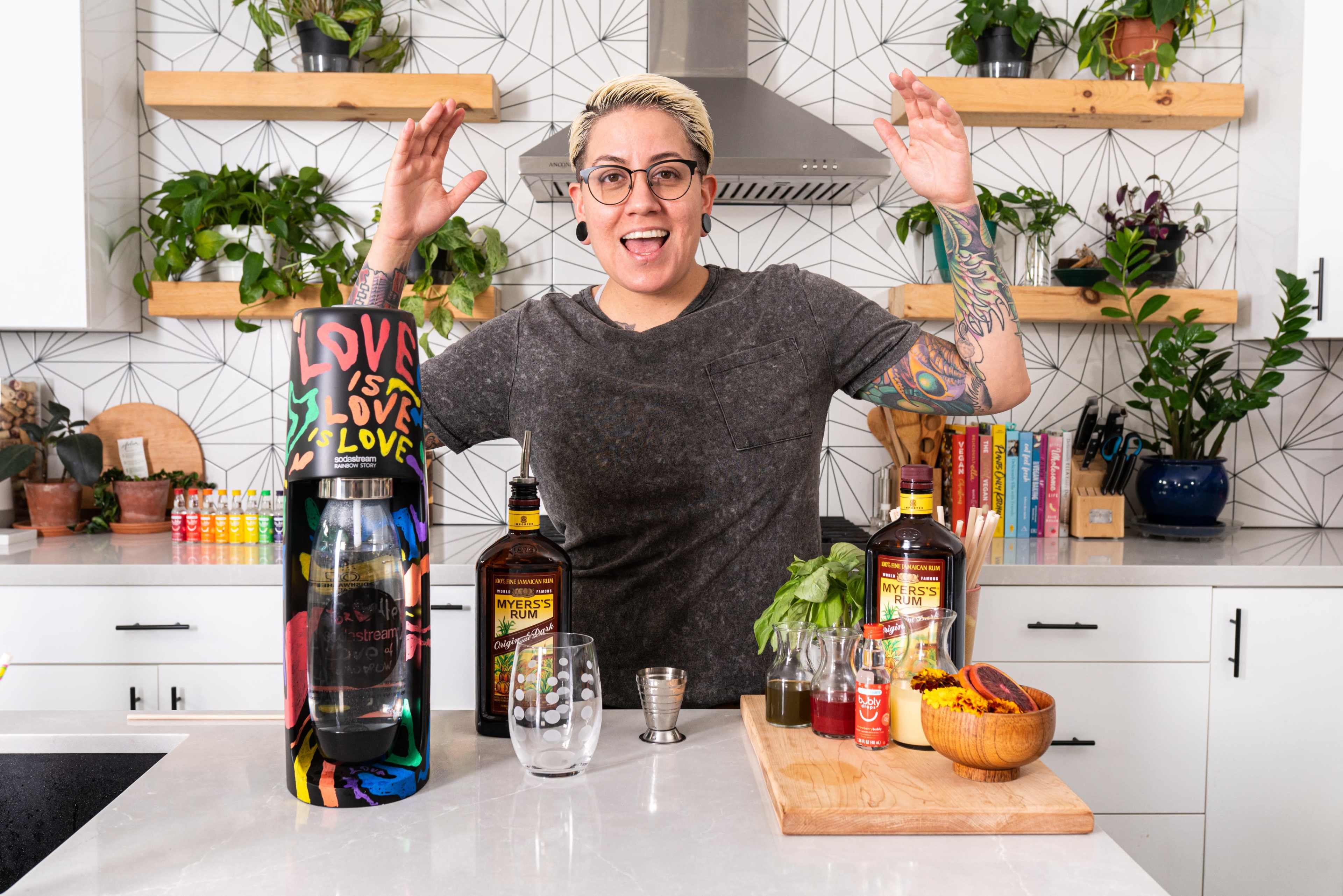 A person with short hair and tattoos stands behind a kitchen counter with various bottles and ingredients for making cocktails, surrounded by potted plants.