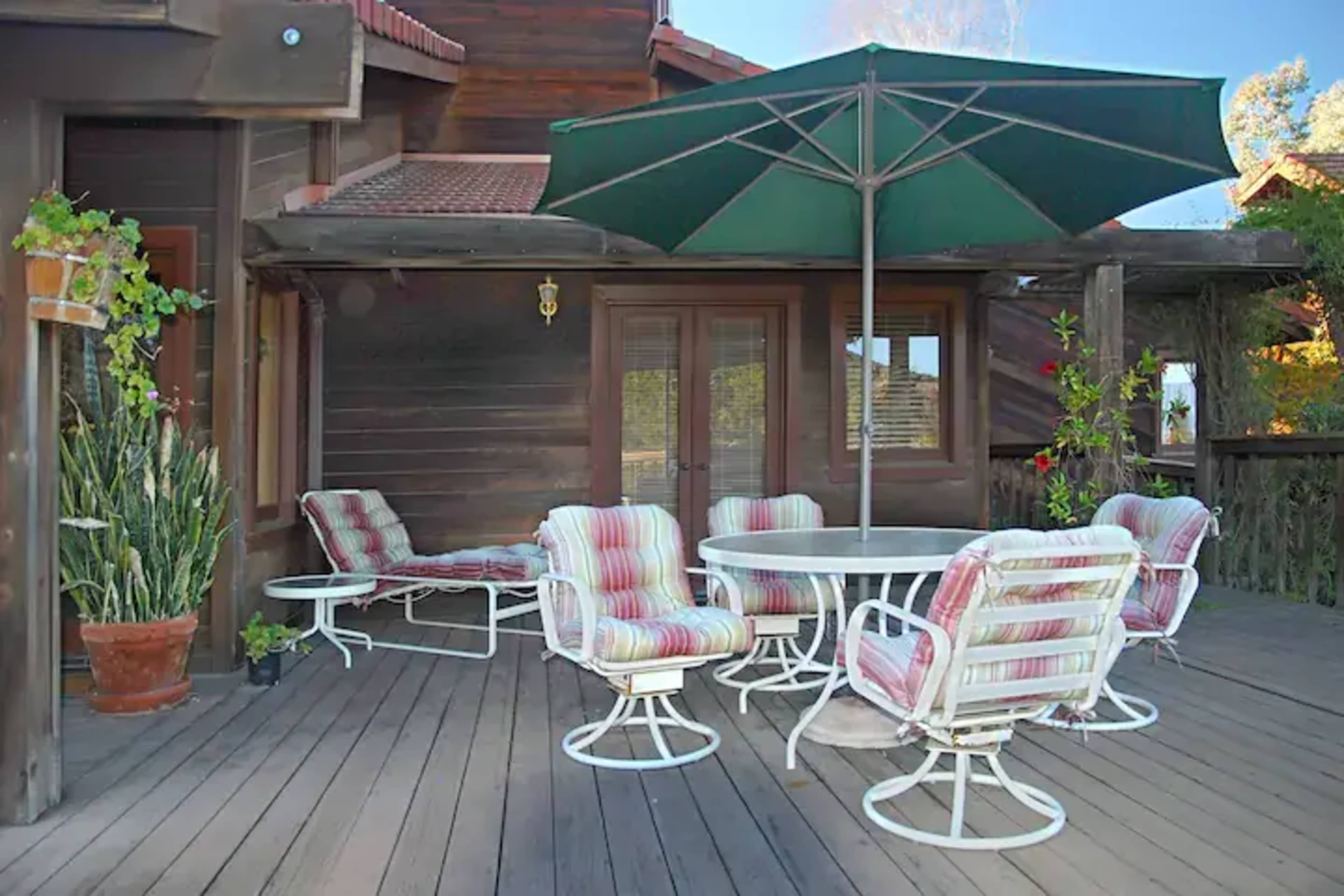 A patio features a round table surrounded by six cushioned chairs and a large green umbrella, with a lounge chair positioned nearby.