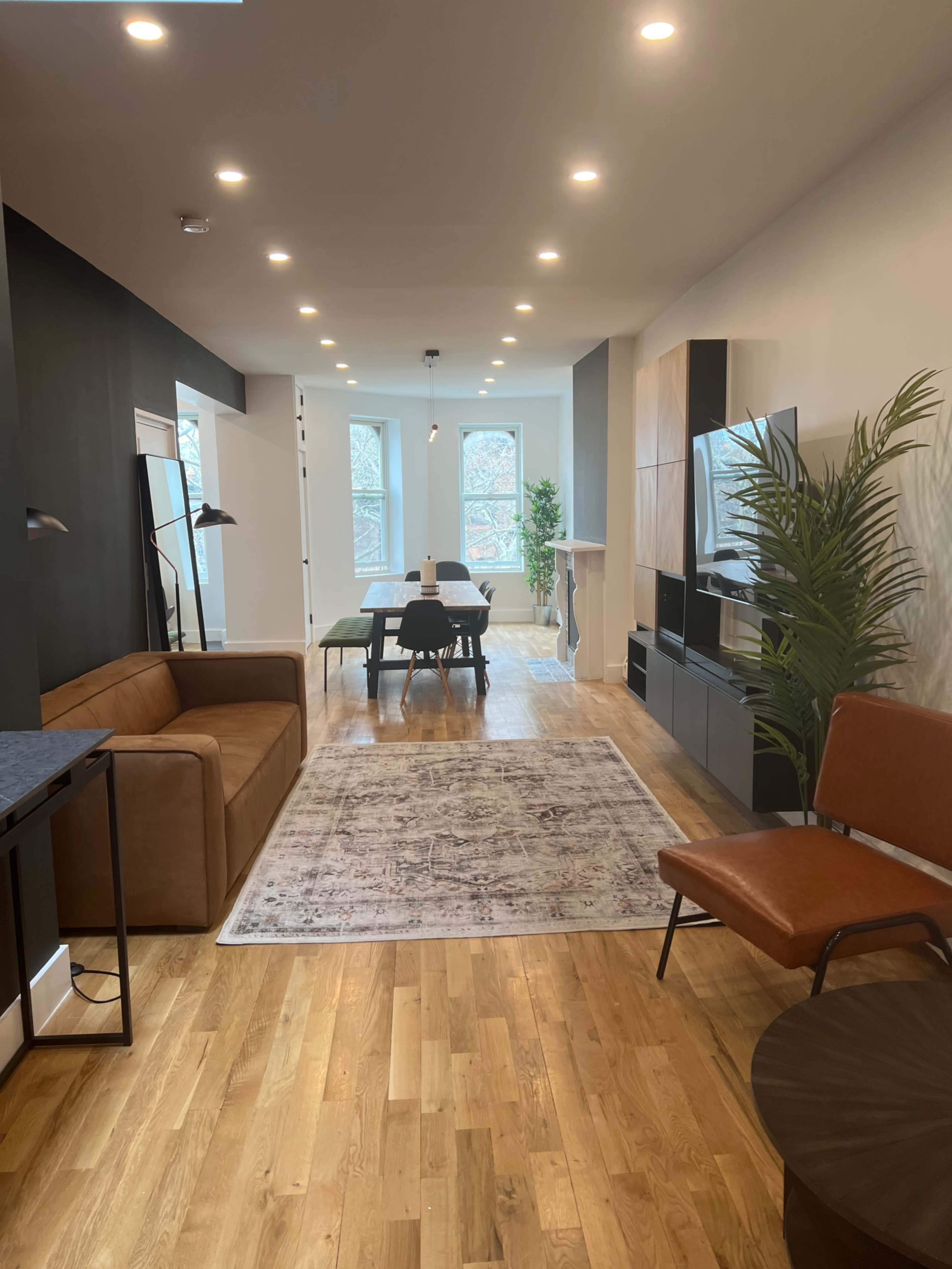 The image shows a modern living space featuring a brown sofa, a dining table, and large windows, with a light wood floor and indoor plants.