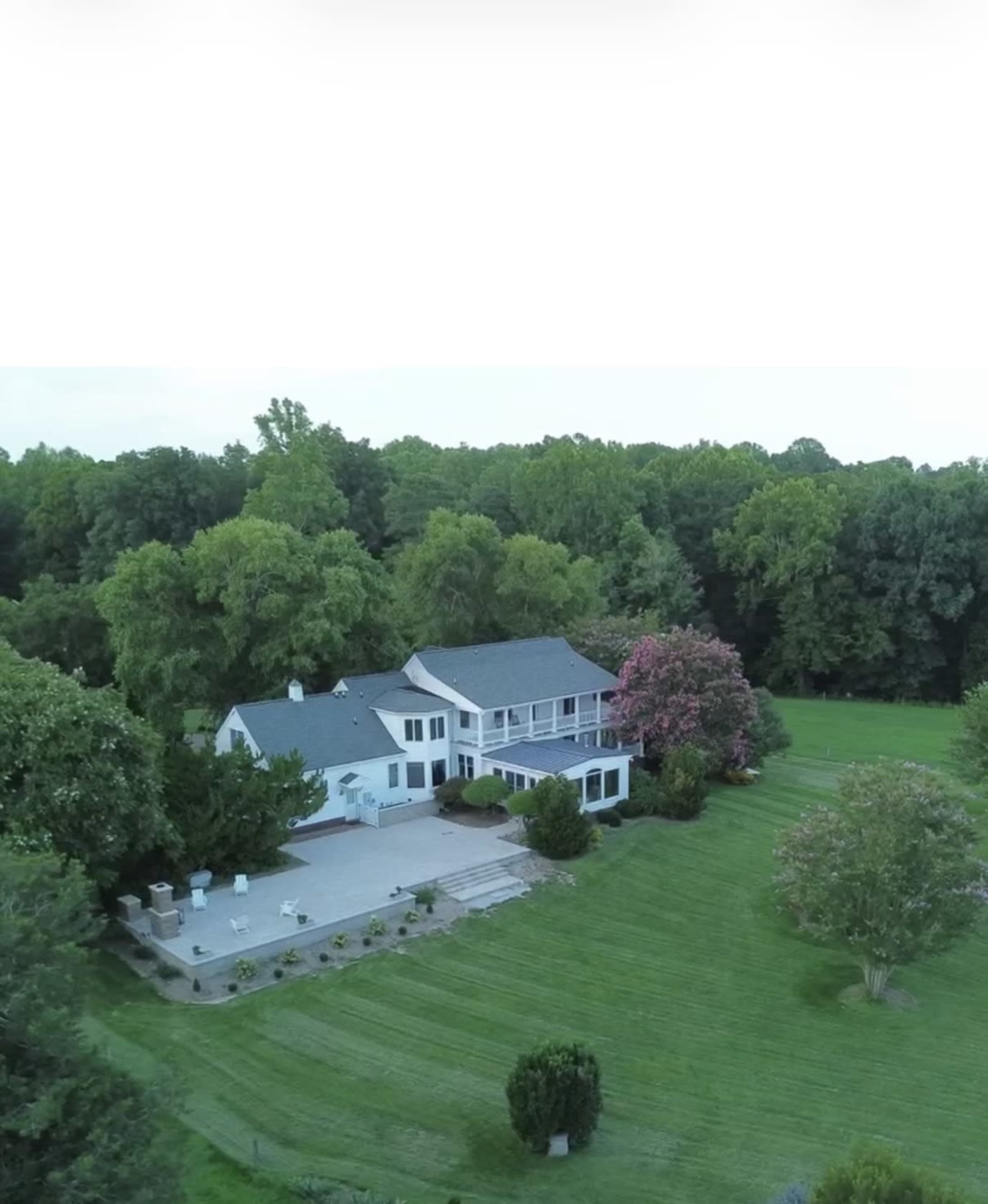 A large, two-story house is surrounded by lush greenery and features a patio area with stone pathways.