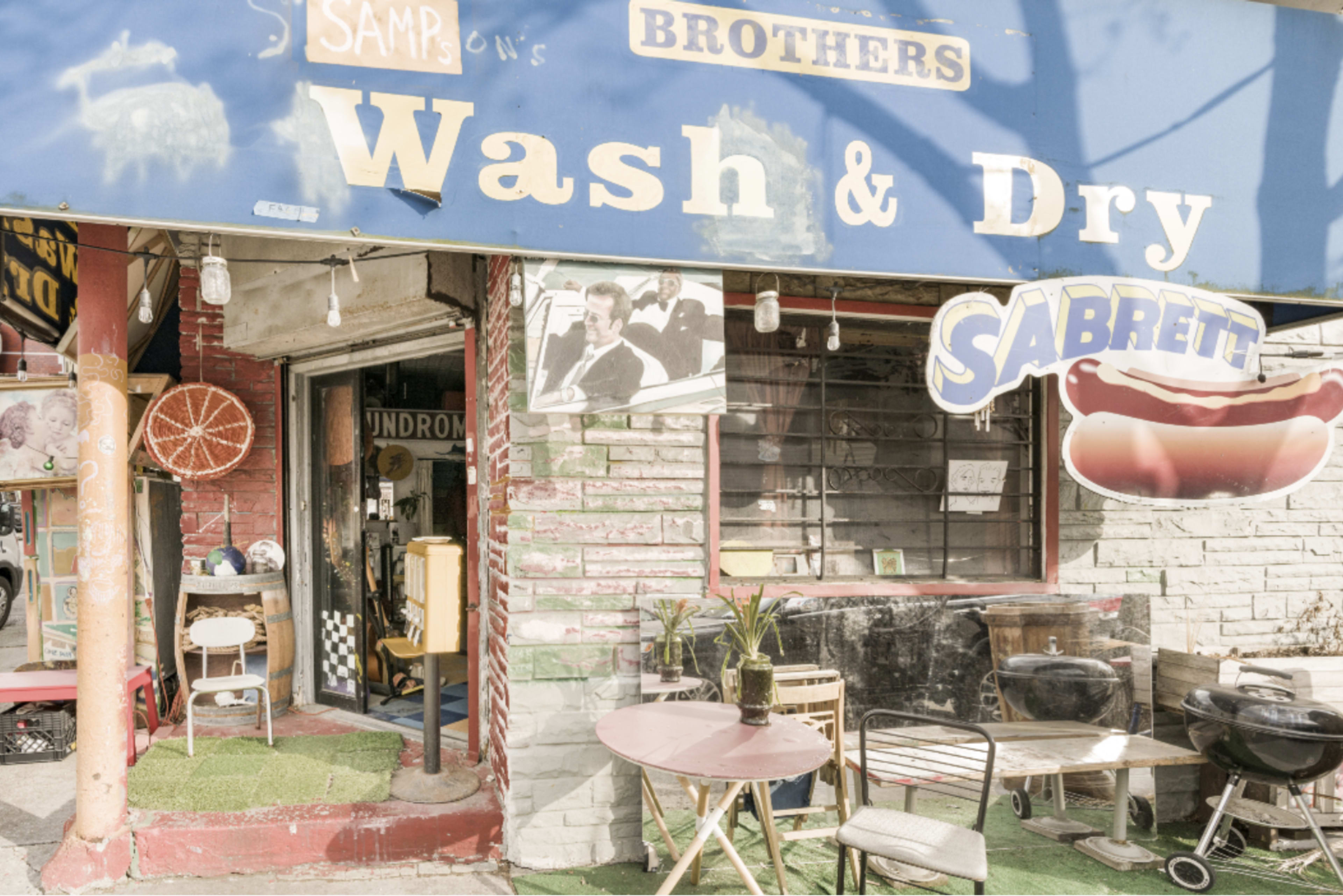 The image shows a laundromat with a blue awning displaying "Wash & Dry," alongside outdoor seating and a hot dog sign.