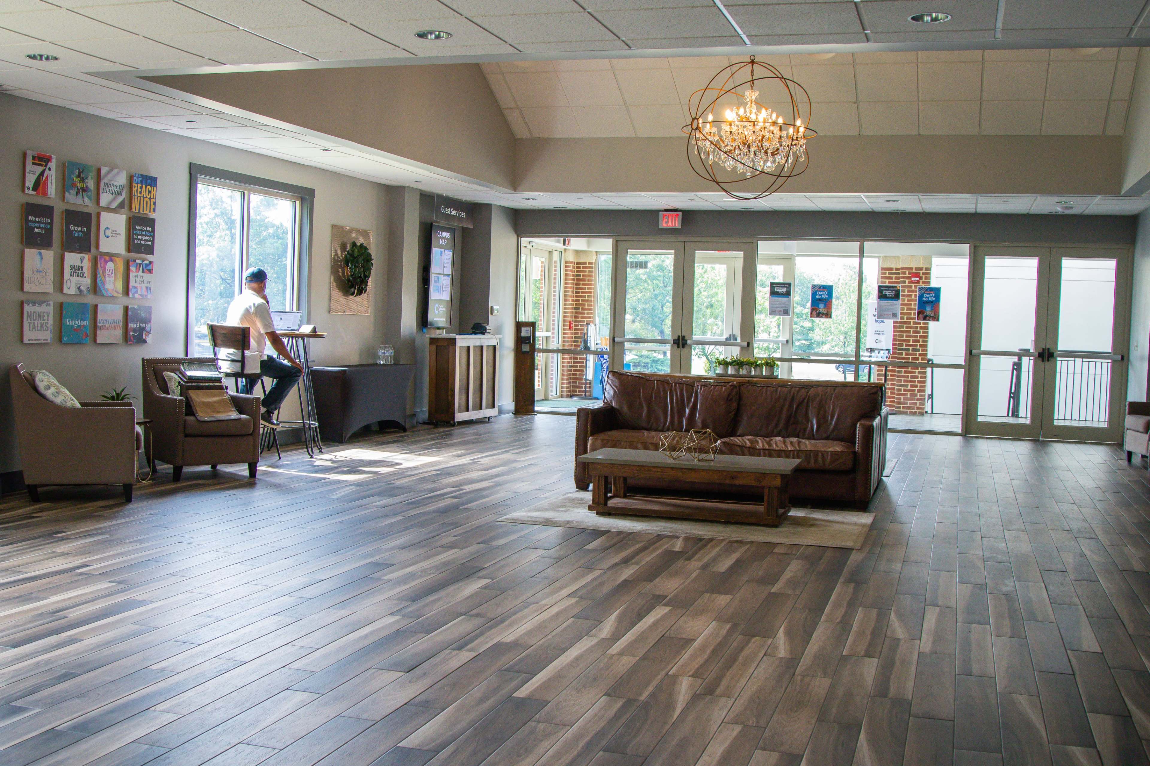 A spacious lobby features a brown leather couch, a coffee table, and a person working at a desk near large windows.