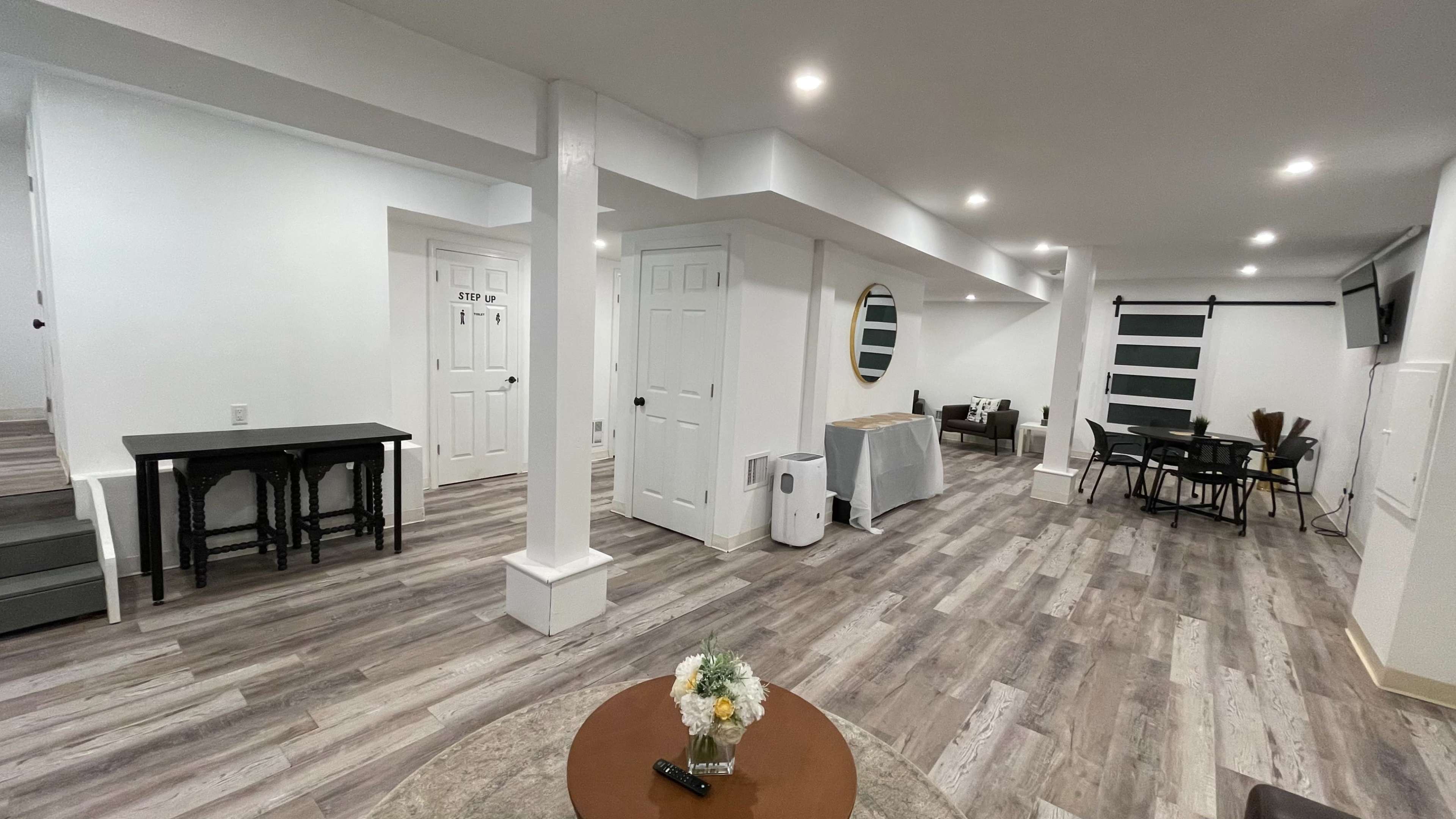 A well-lit, modern basement living space features a sitting area, dining table, and a kitchenette with a large mirror on the wall.
