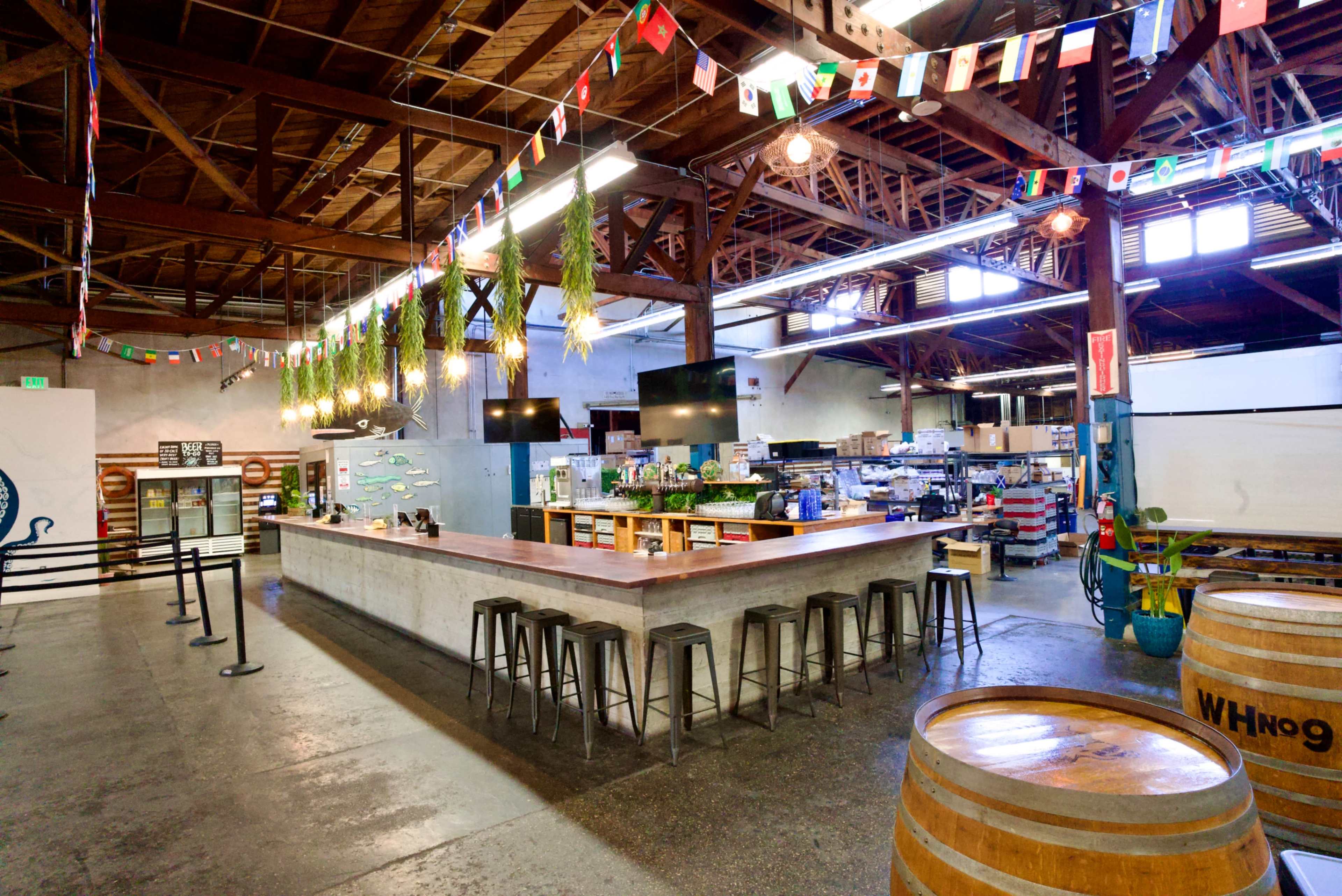 The image shows an industrial-style food market with a long bar area, wooden barrels, and colorful flags hanging from the ceiling.