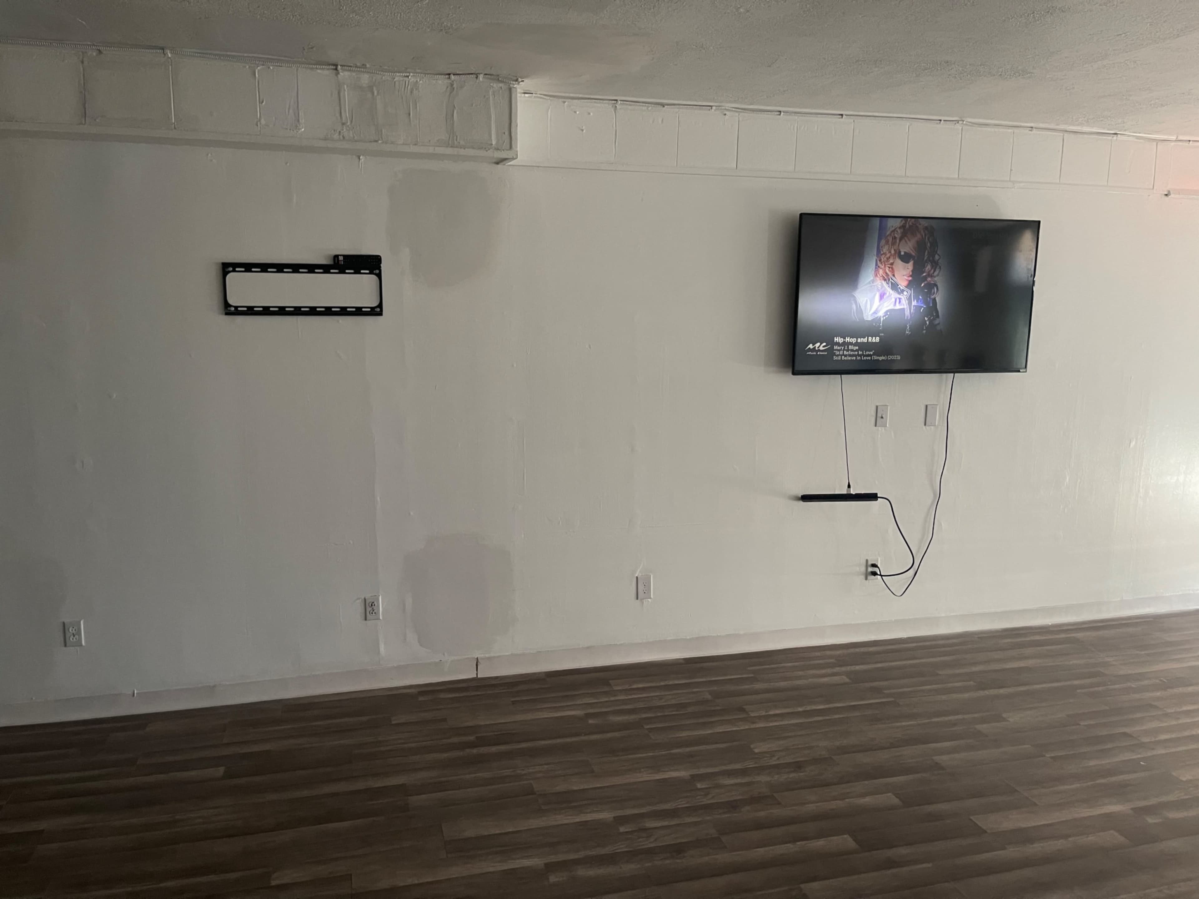 A sparsely furnished room with a mounted television on one wall and a blank section of wall next to it, featuring wooden flooring.